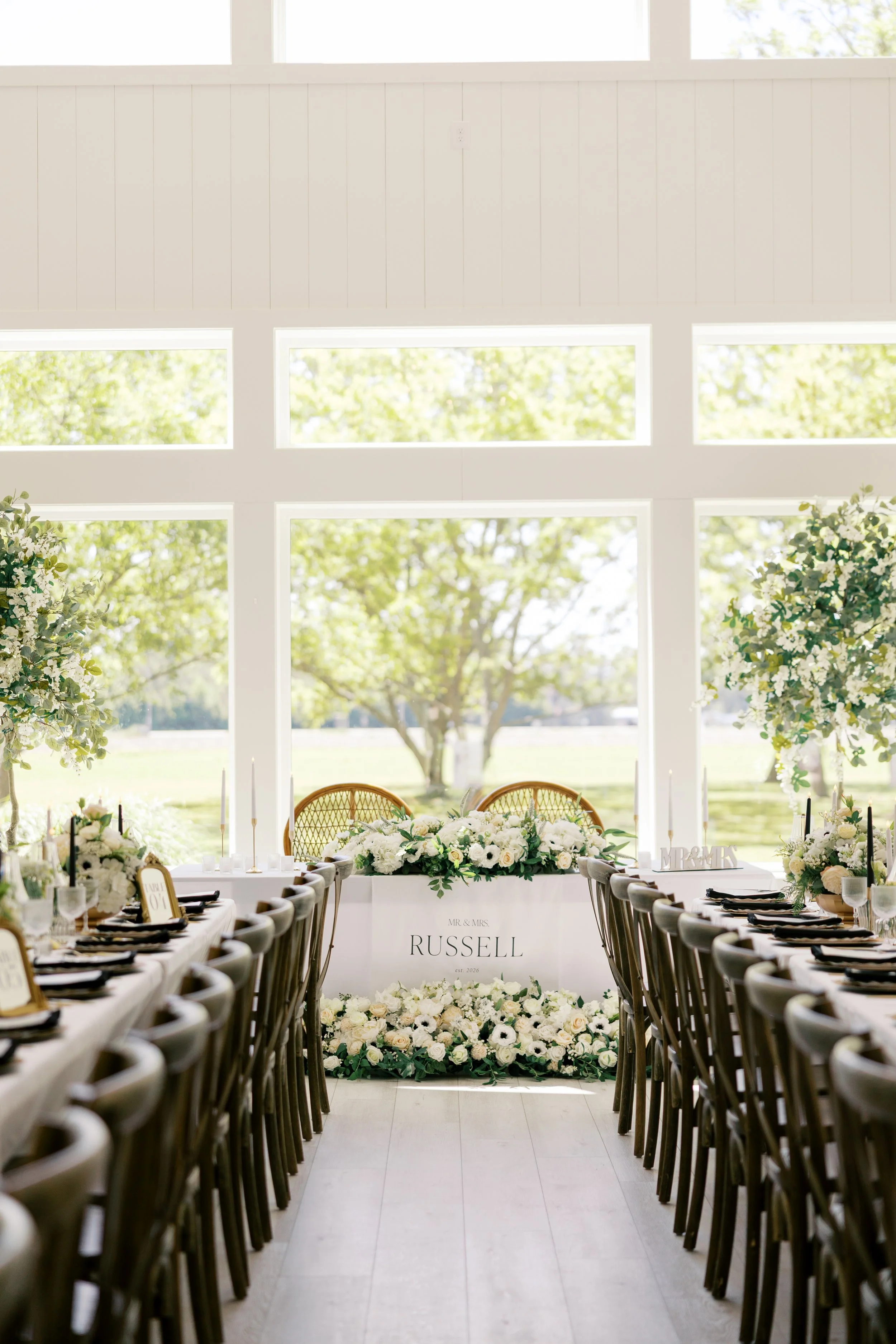Wedding reception setup with long tables decorated with white flowers, greenery, and candles, with a head table featuring a large floral arrangement and a sign reading "Mr. & Mrs. Russell" in a bright room with large windows showing trees outside.