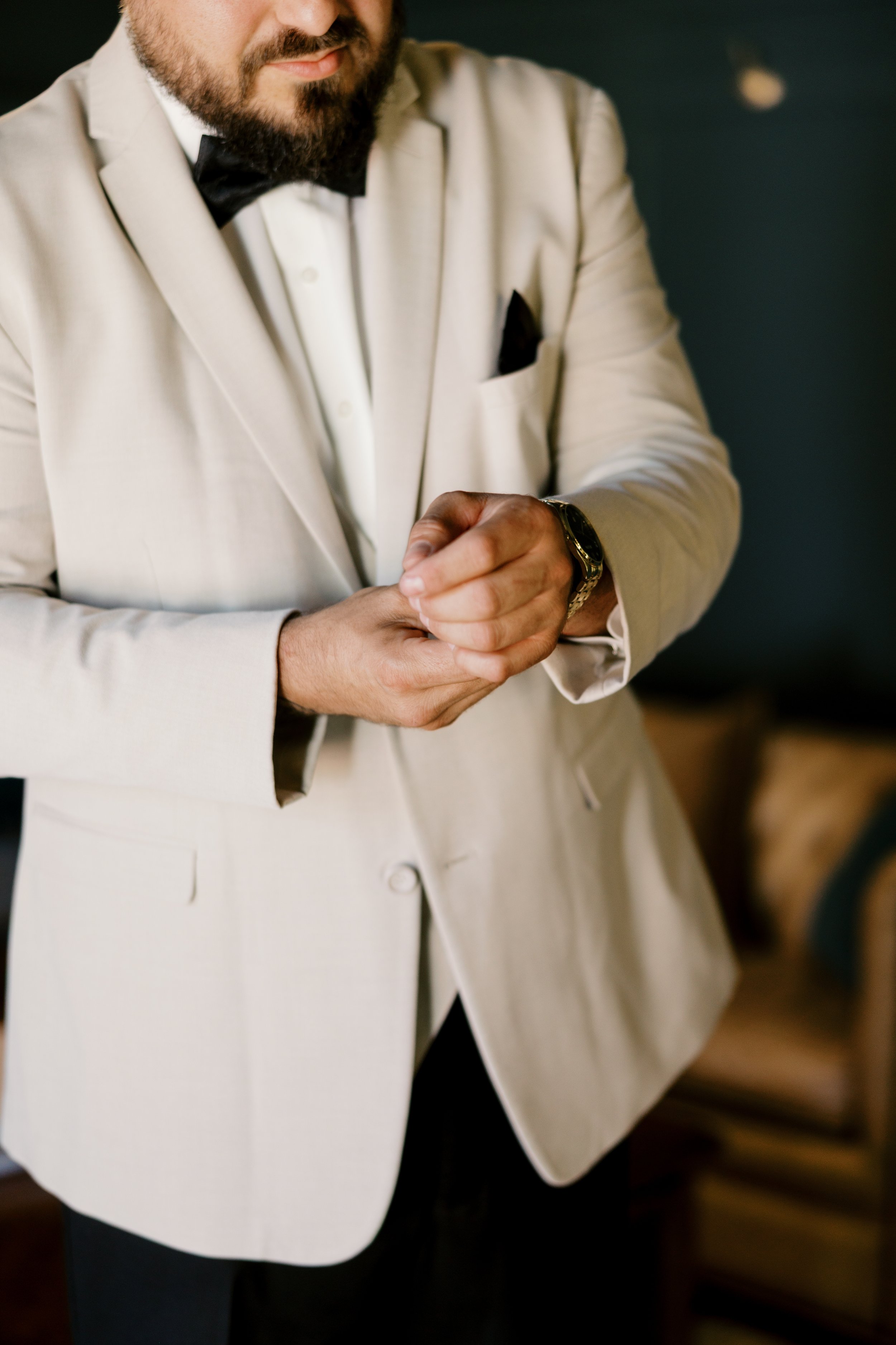Close-up of a man wearing a white tuxedo, black bow tie, and a wristwatch, adjusting his cufflink.