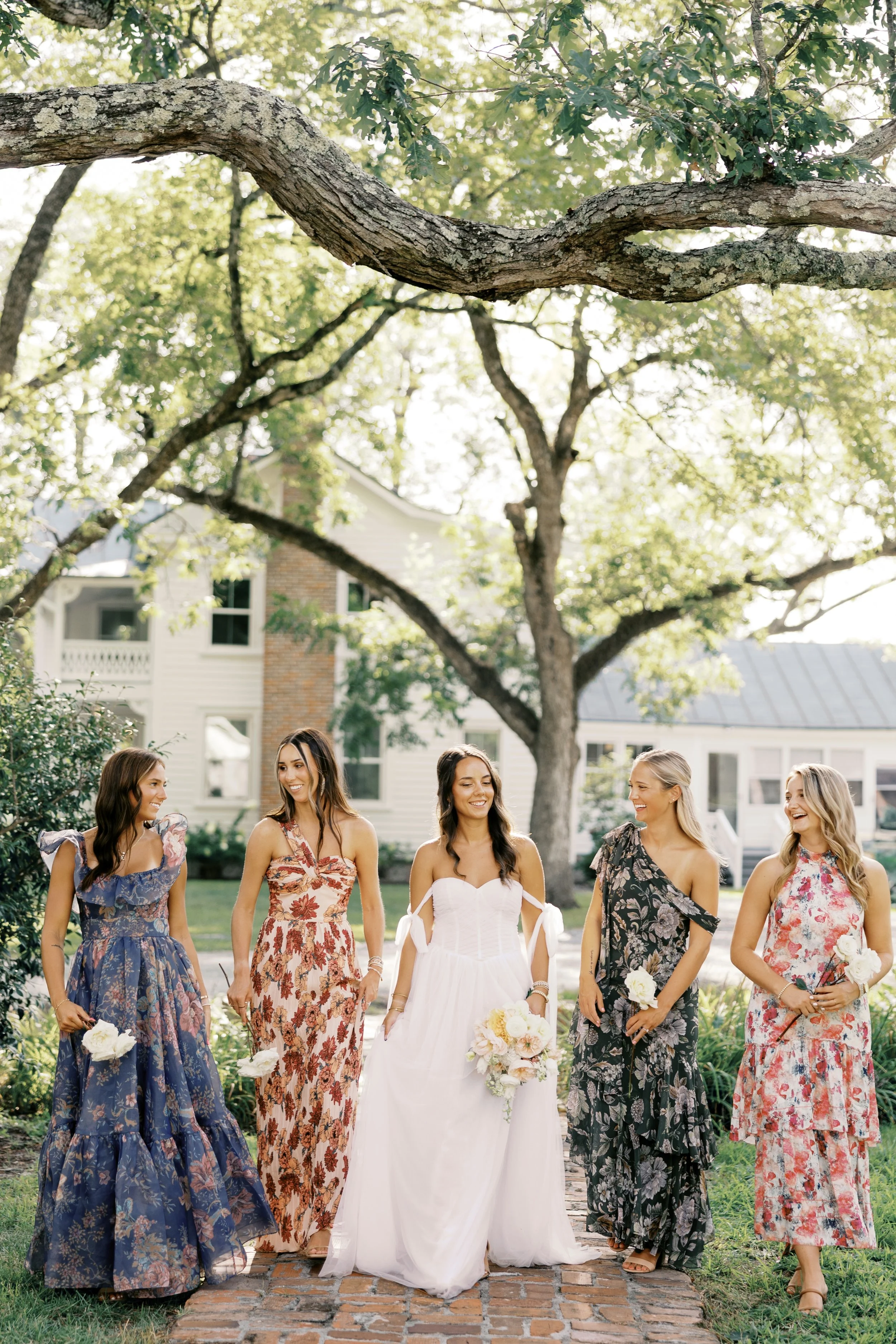 Group of six women in colorful dresses walking outdoors in a garden during a wedding celebration.