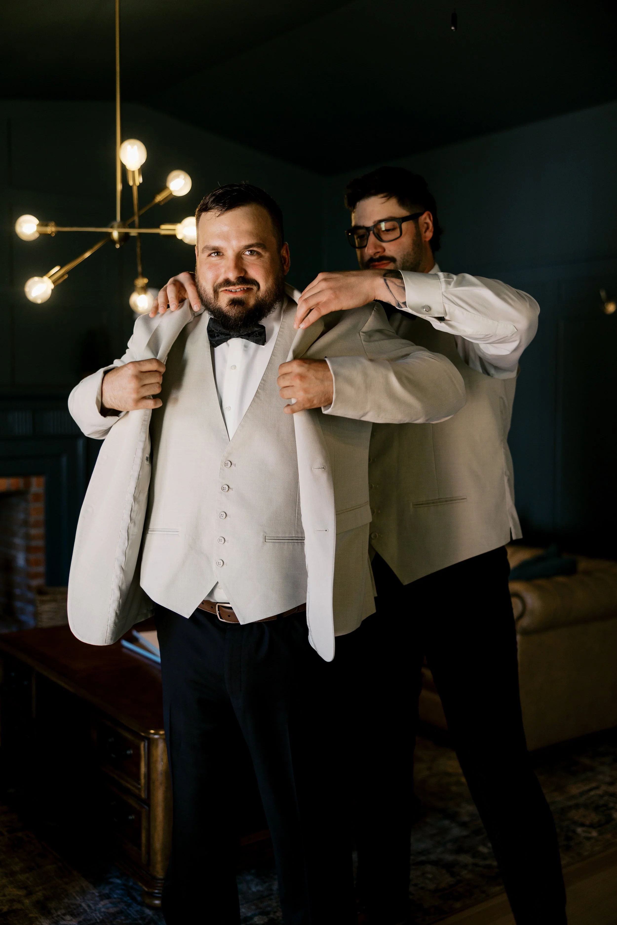 Two men in formal attire, one helping the other adjust his suit jacket, in a dimly lit room with a modern chandelier and a dark-colored wall.