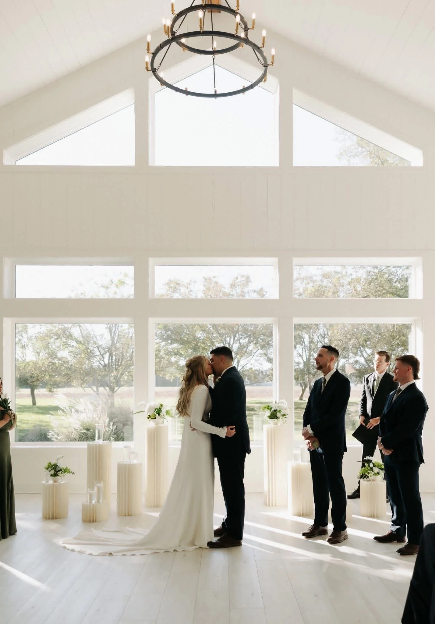 A bride and groom kissing during their wedding ceremony in a bright, modern venue with large window panels and a view of trees outside, with groomsmen standing to the right of the couple.