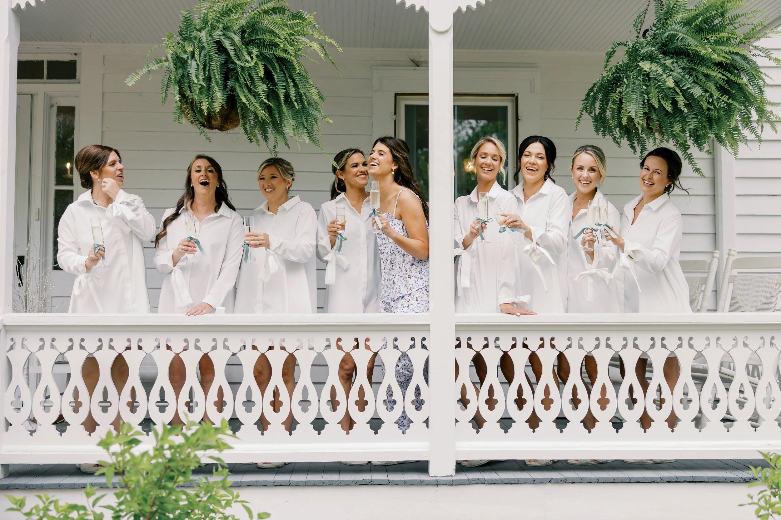 A group of women on a porch celebrating with champagne glasses, dressed in white and floral dresses, with hanging plants and green foliage for decor.