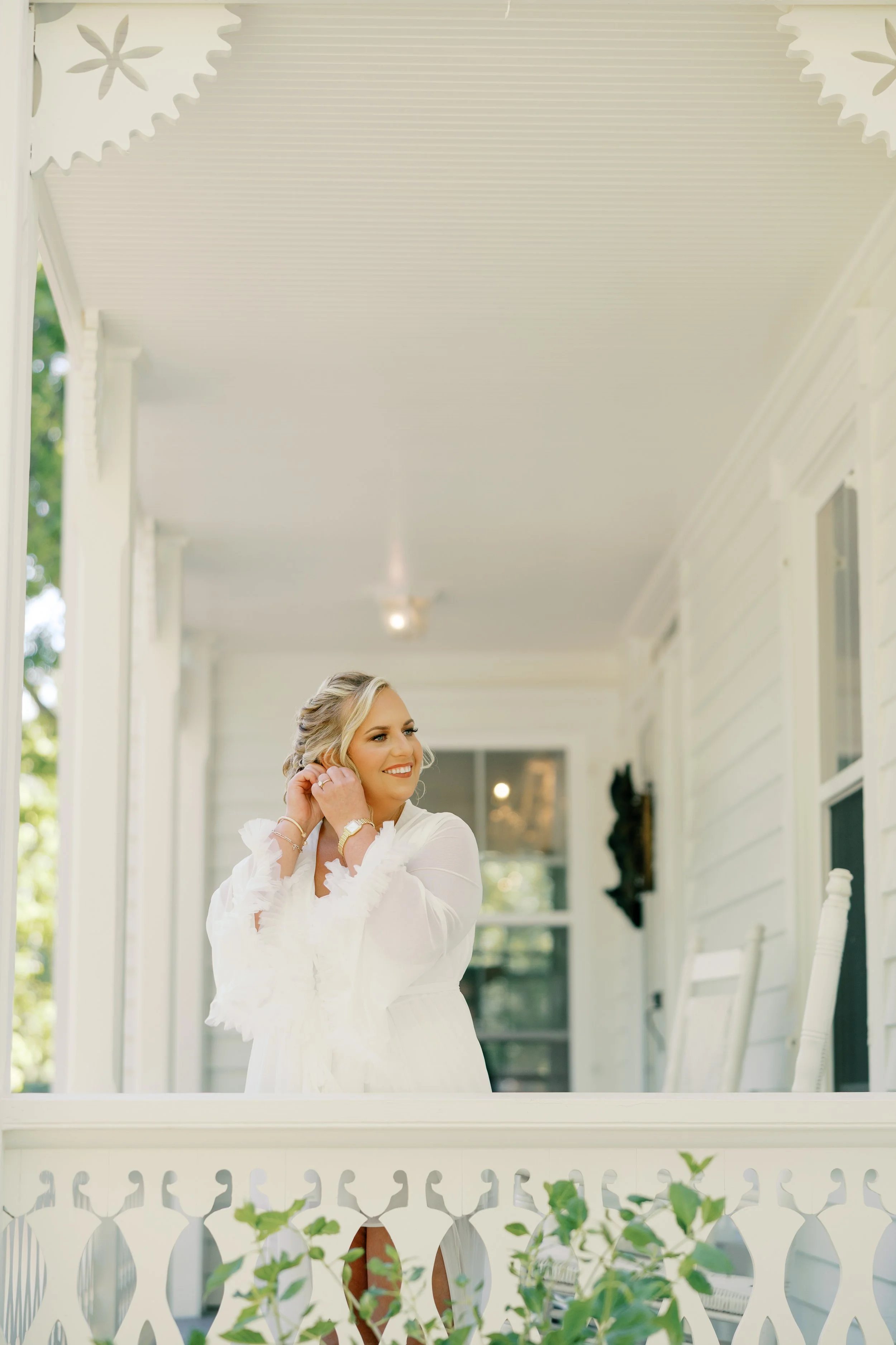 A woman in a white dress, smiling, standing on a porch, adjusting her earring, with the porch's white railing and house exterior visible in the background.