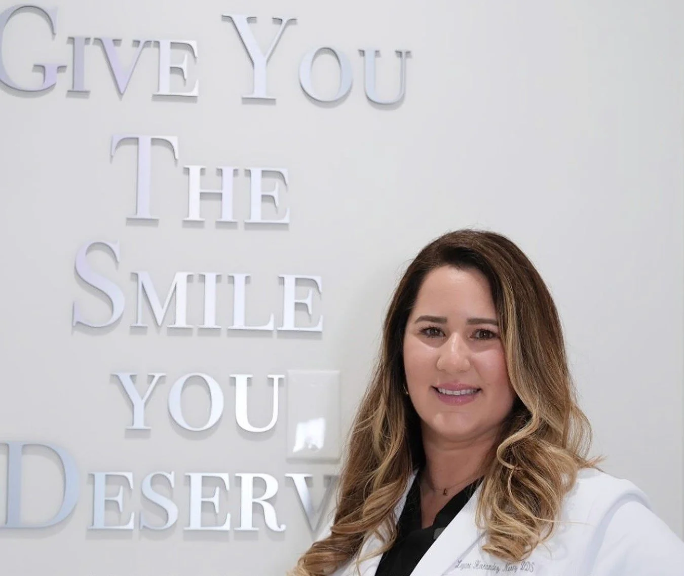 A woman with long, wavy brown hair smiling in front of a white wall with 3D white letters that say "GIVE YOU THE SMILE YOU DESERVE"