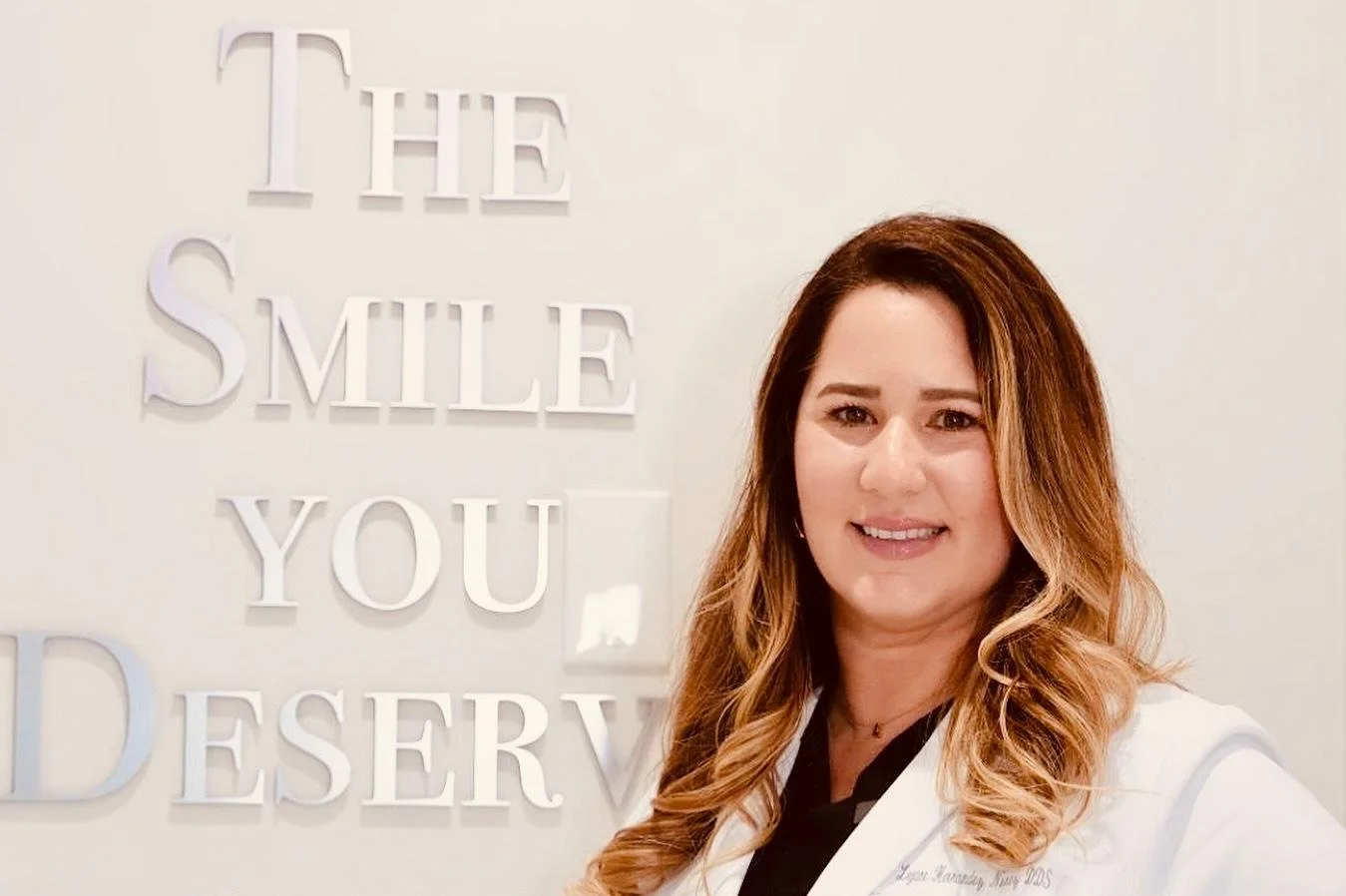 A woman with long brown wavy hair smiling, standing in front of a wall with the text "The Smile You Deserve" on it.