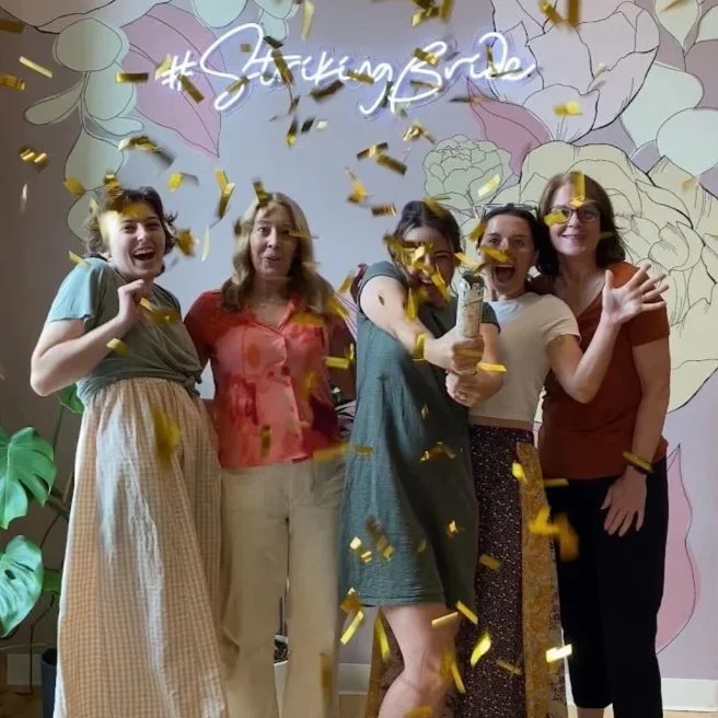 Five women celebrating saying "yes to the dress" with gold confetti, a glowing sign saying '#strikingbride', and floral mural in the background.