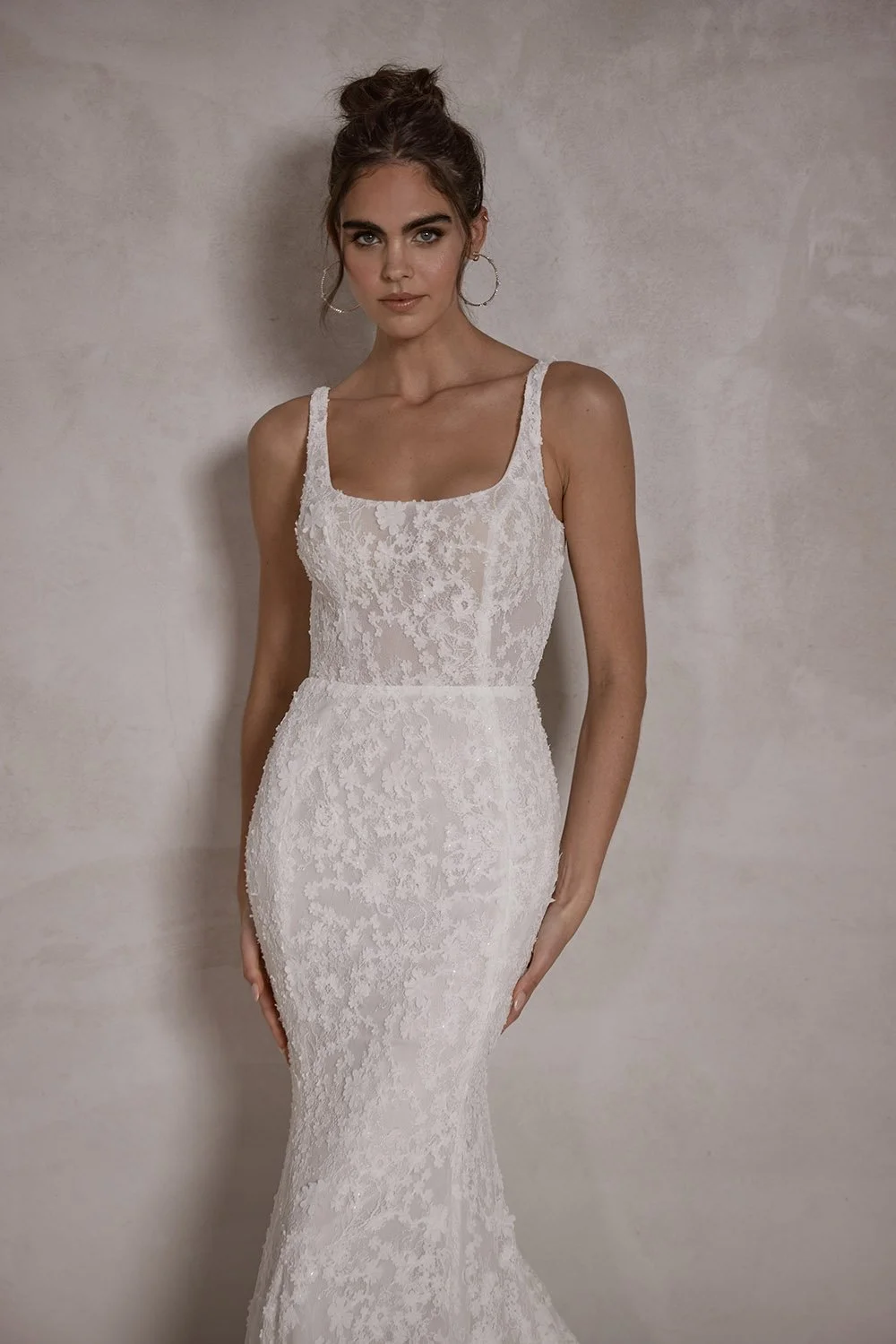 A woman wearing a white lace bridal gown with a square neckline and fitted silhouette, standing against a plain light-colored wall.