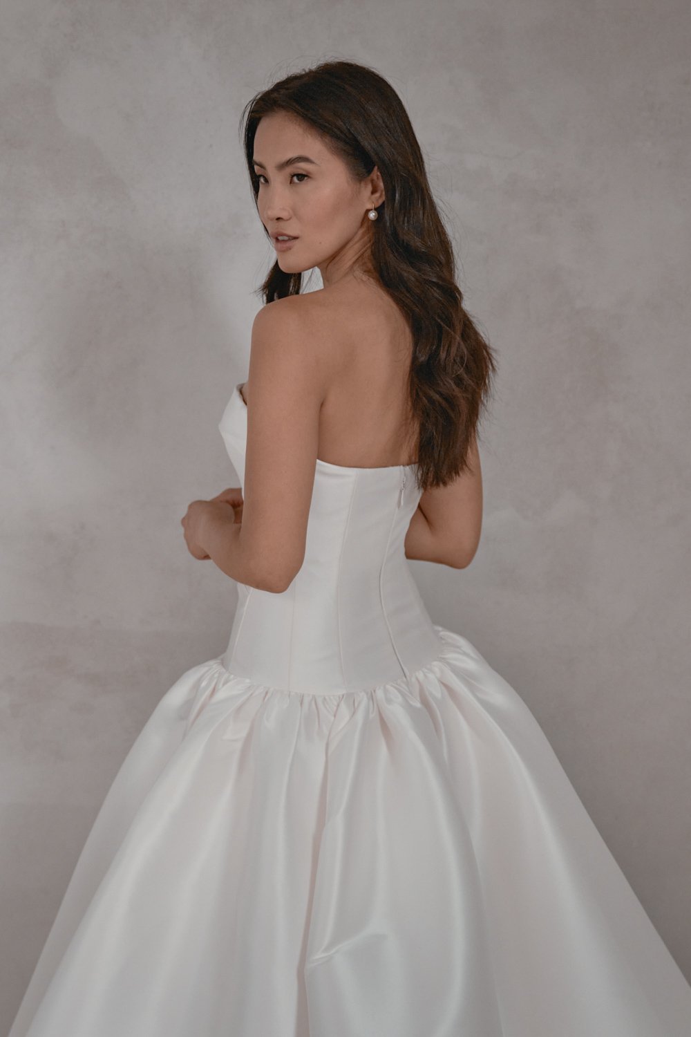 A woman in a strapless white wedding dress with a fitted bodice and a full, gathered skirt, standing against a plain background, looking over her shoulder.