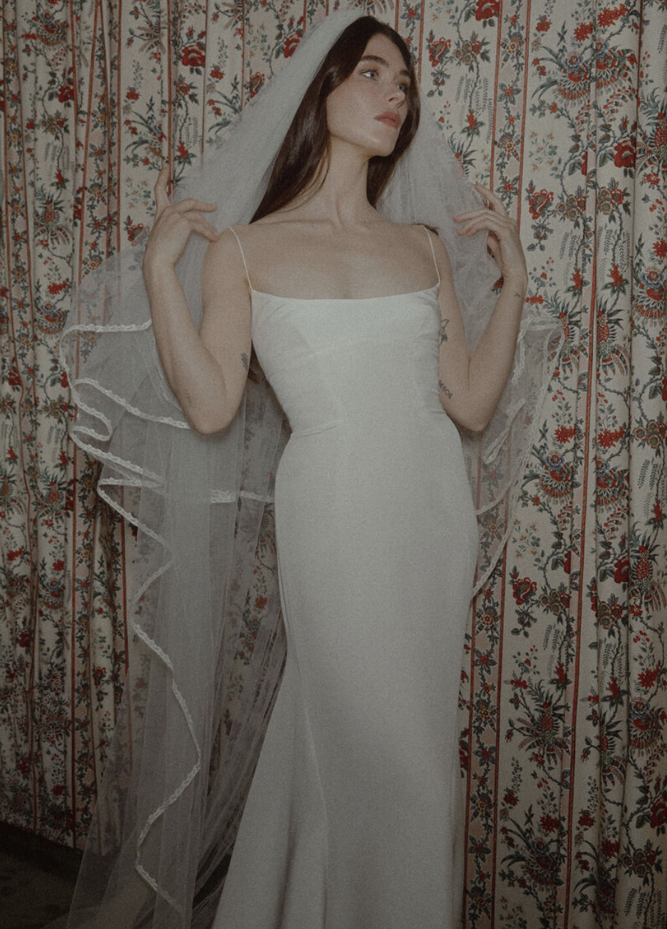A woman in a white wedding dress with thin straps, standing in front of a floral patterned curtain, holding a veil.