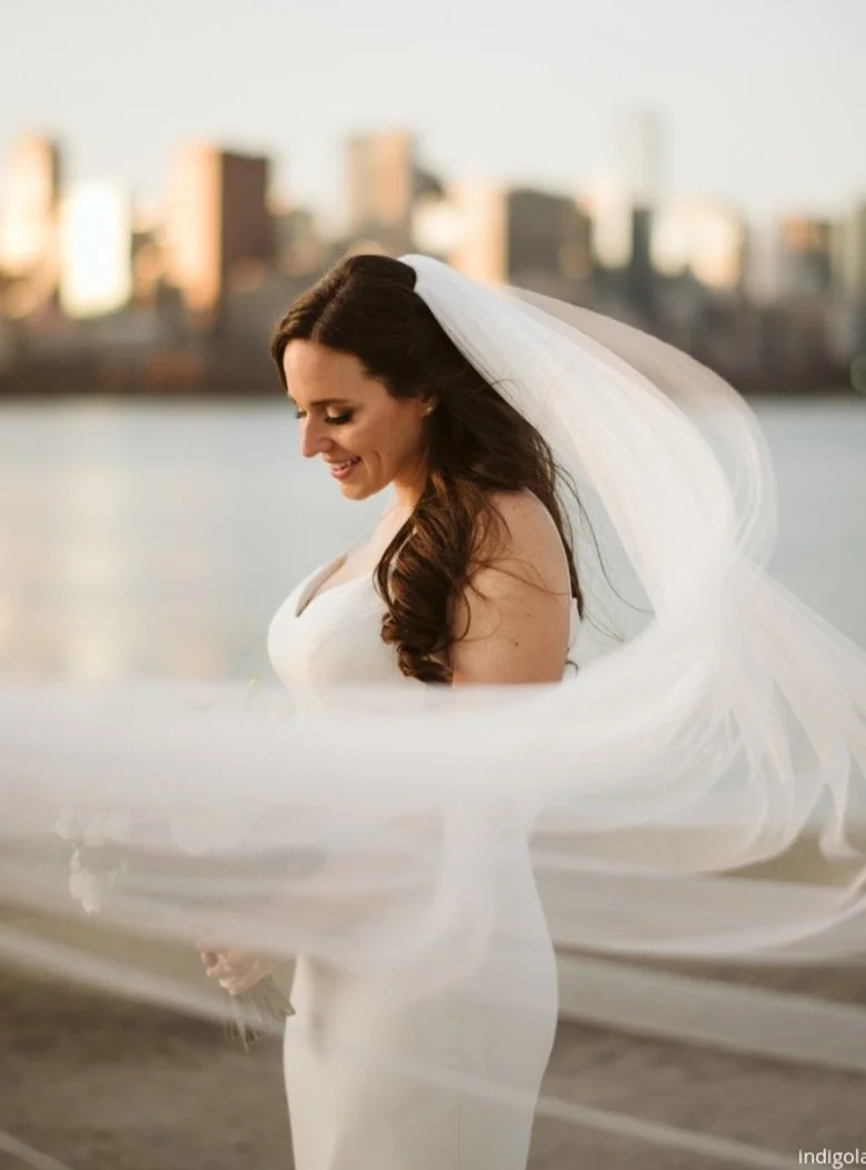 A woman in a white wedding gown with a veil, standing outdoors near a body of water with a city skyline in the background, smiling and looking down.