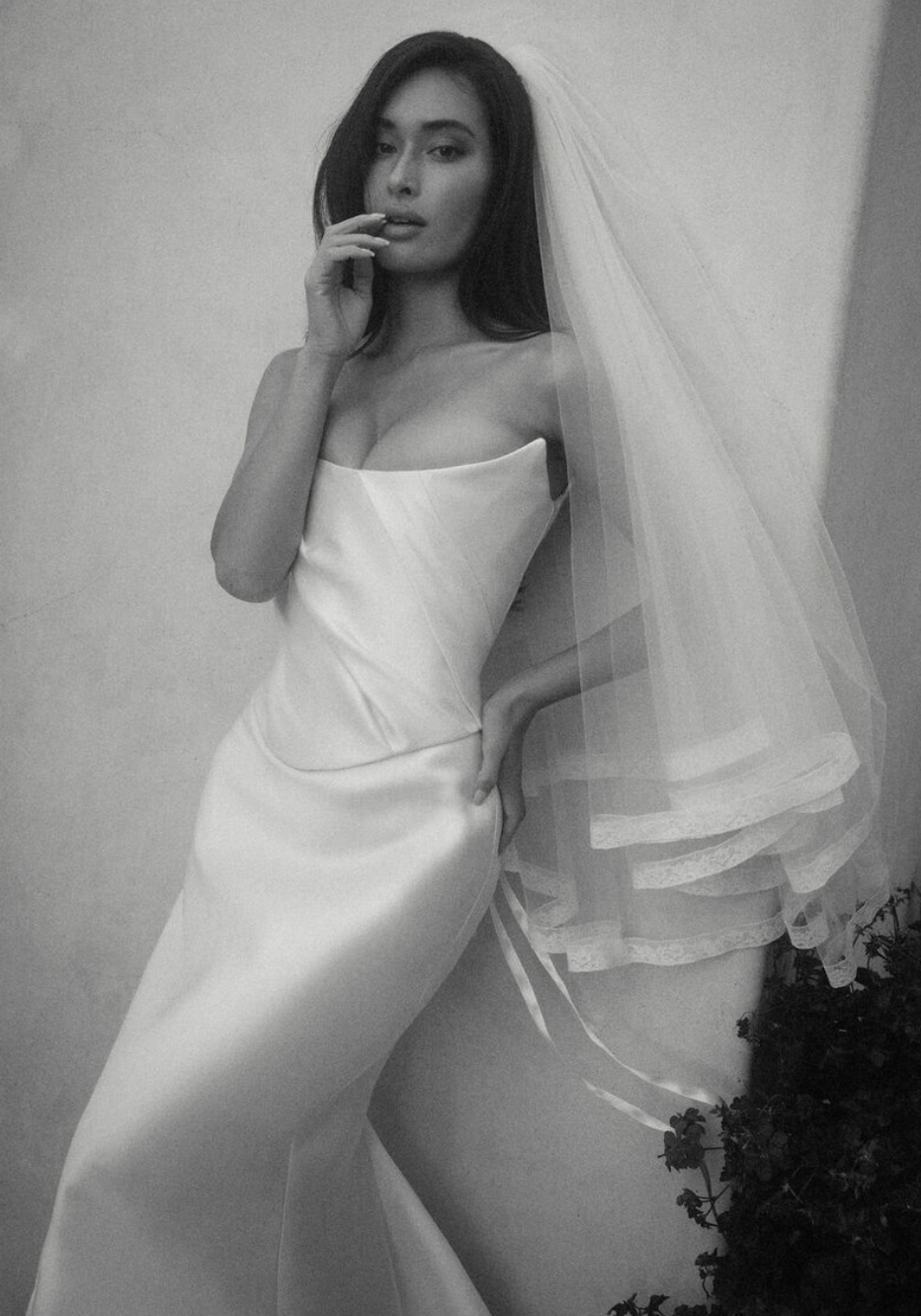 Black and white photo of a woman in a strapless gown with a veil, posing with one hand on her hip and the other near her face.
