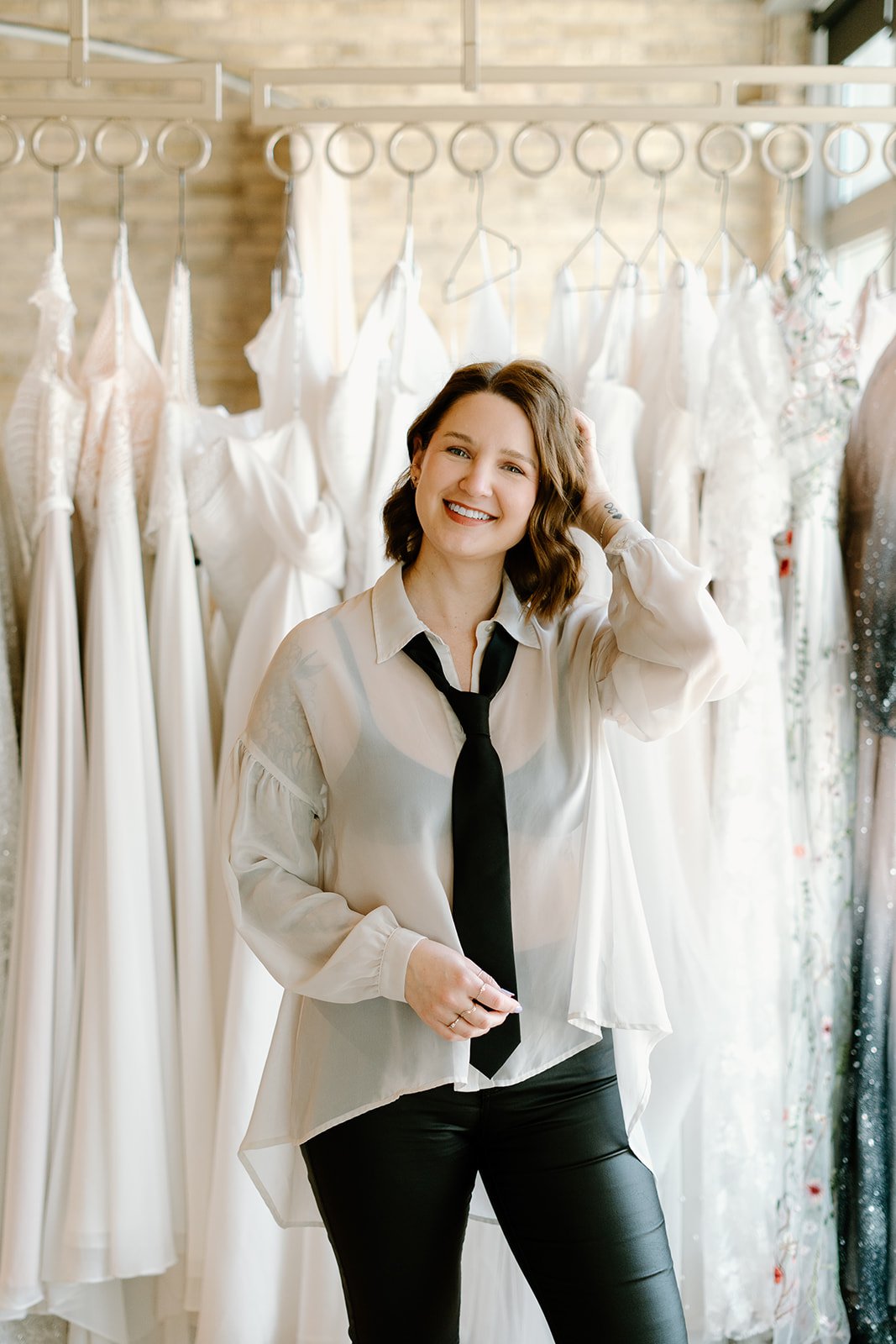 A woman with wavy brown hair smiling and wearing a white shirt and black tie, standing in front of a display of white wedding dresses hanging on a rack.