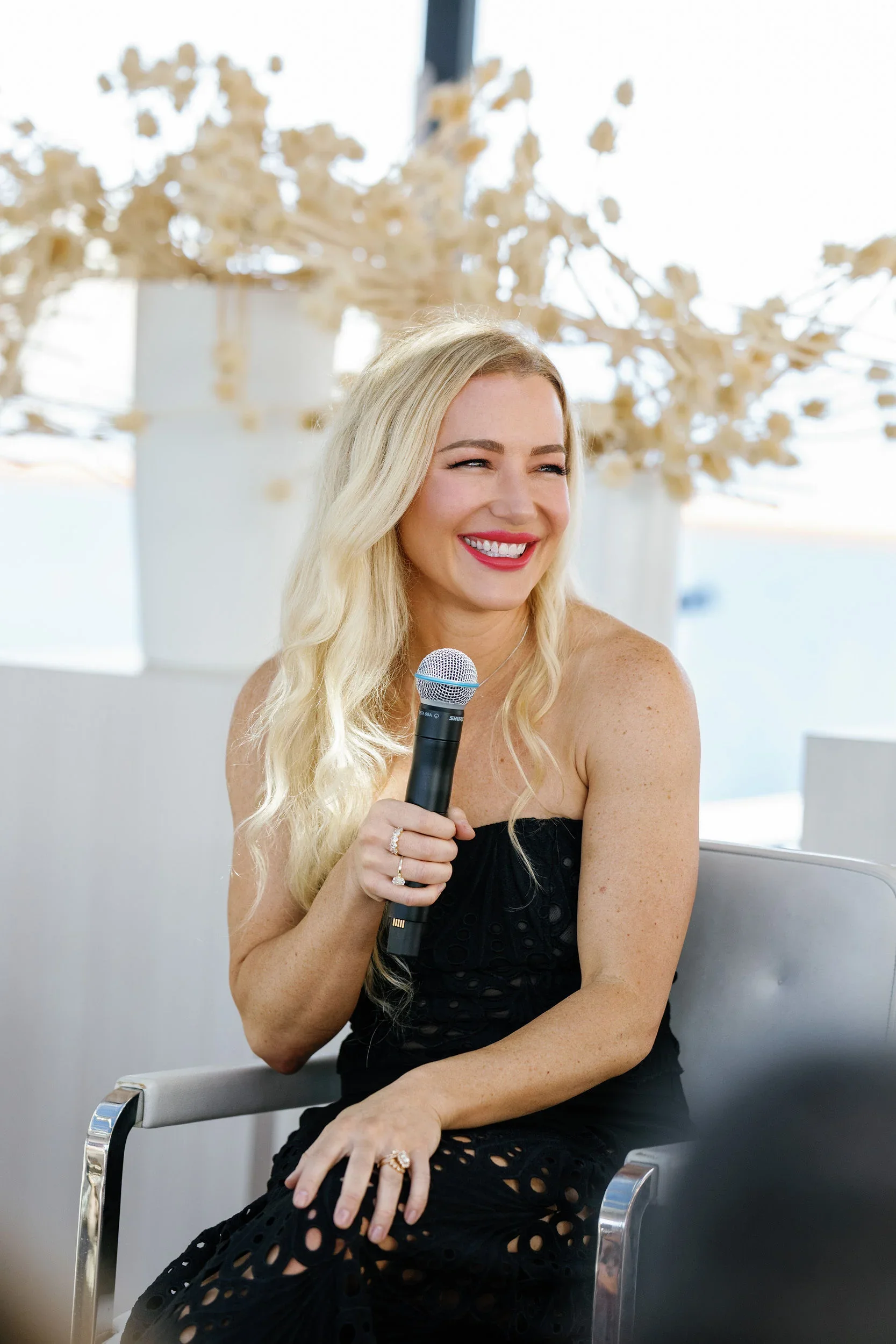 A woman with long blonde hair and a black dress sitting on a white chair, smiling while holding a microphone in her right hand, with a large floral arrangement in the background.