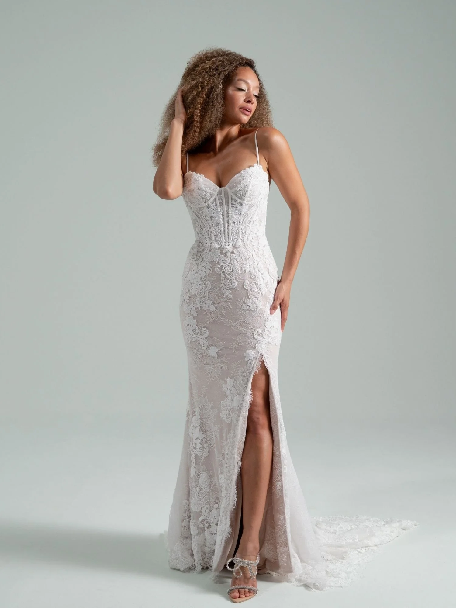 A woman wearing a white lace wedding dress with spaghetti straps, a slit revealing her leg, and high heels, standing against a plain light gray background.