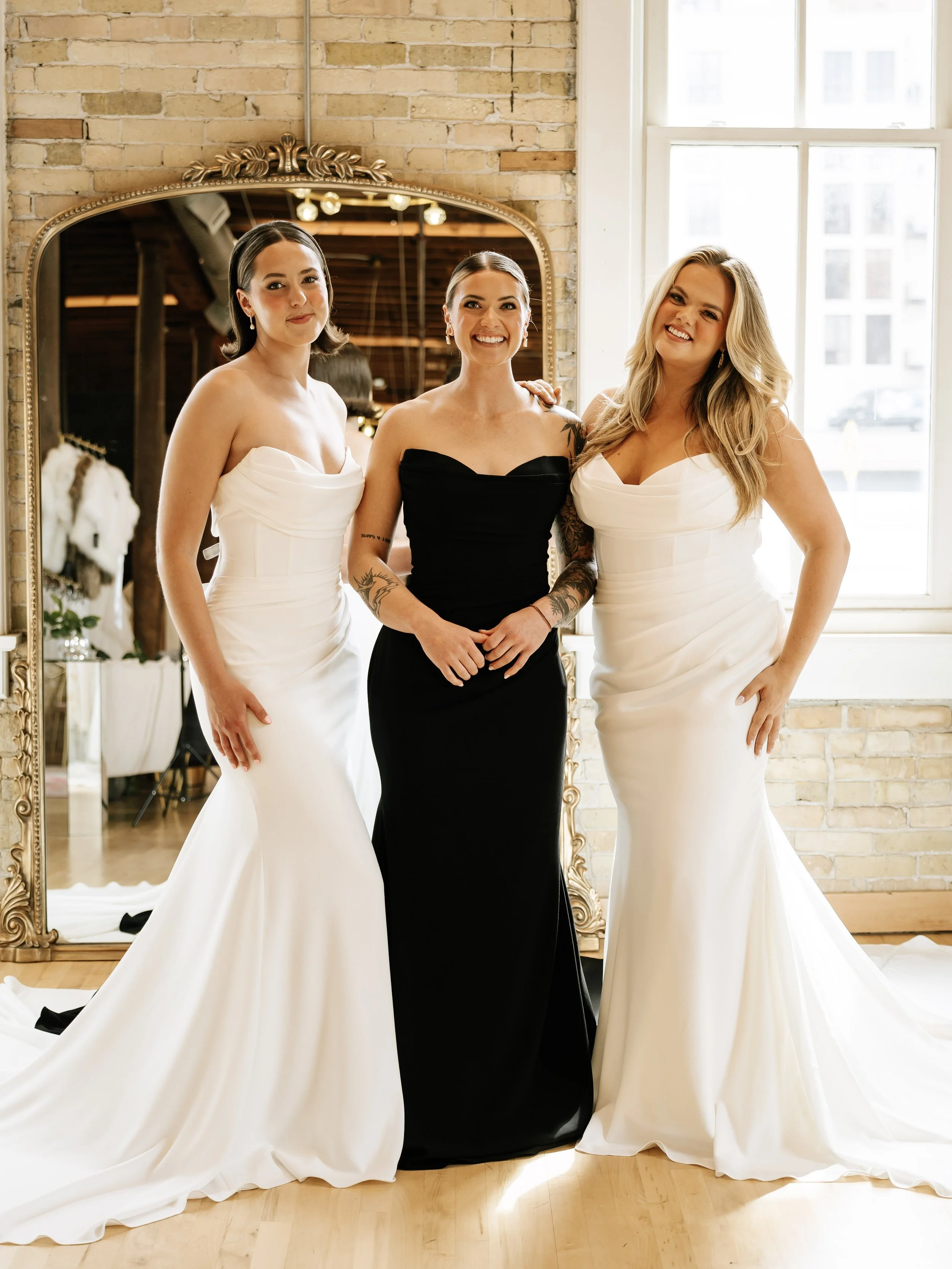 Three women dressed in wedding gowns, one in a black dress, standing in front of a large ornate mirror in a room with a brick wall and large windows. They are smiling and posing together.