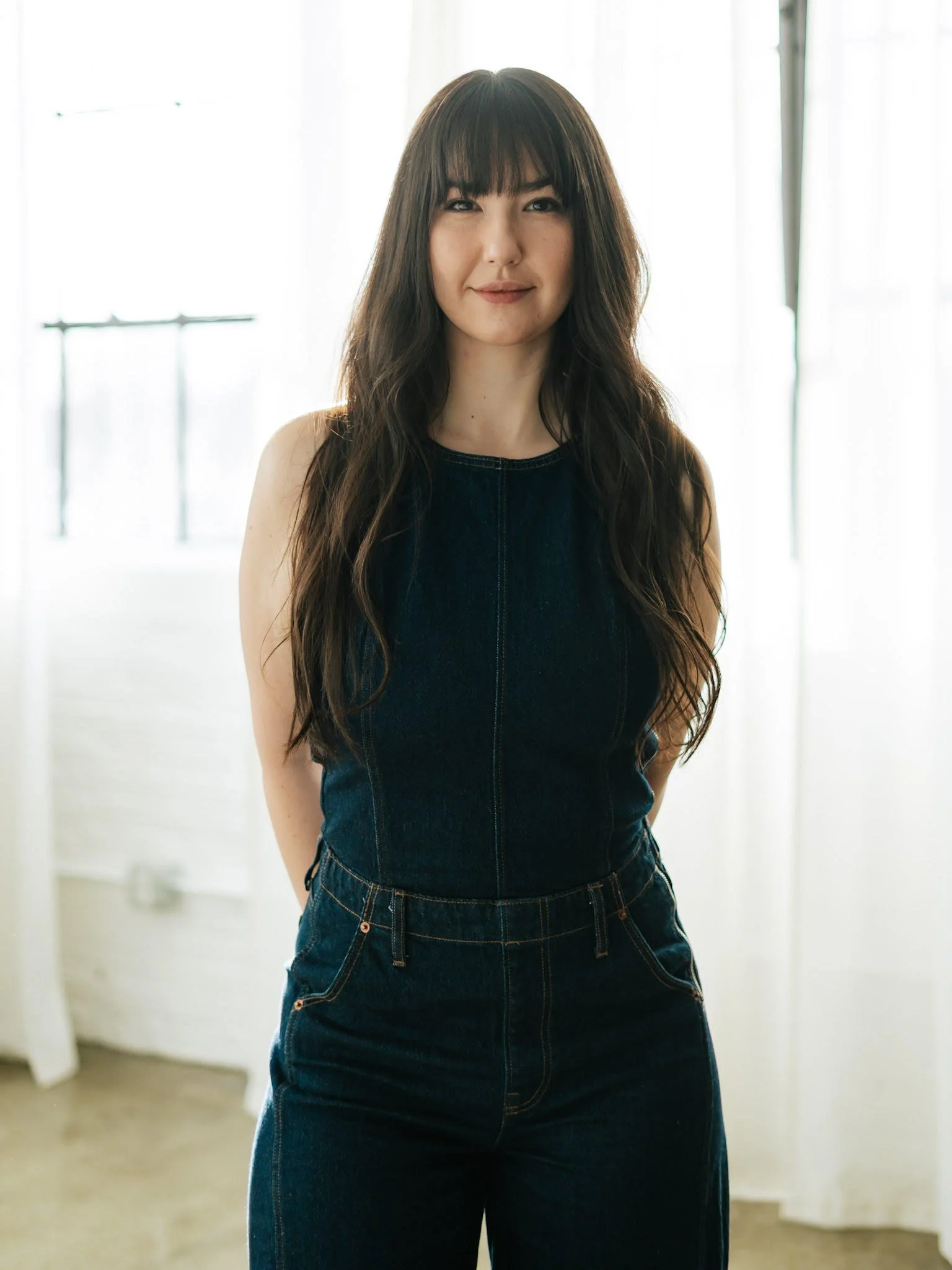 A woman with long dark hair and bangs wearing a sleeveless dark denim dress, standing in a bright room with sheer curtains.