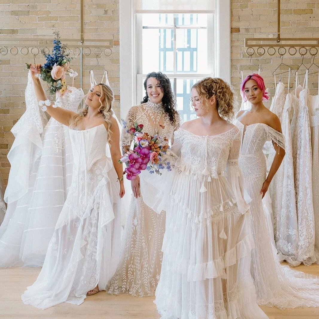 Four women in wedding dresses shopping for bridal gowns in a boutique with windows and brick walls.