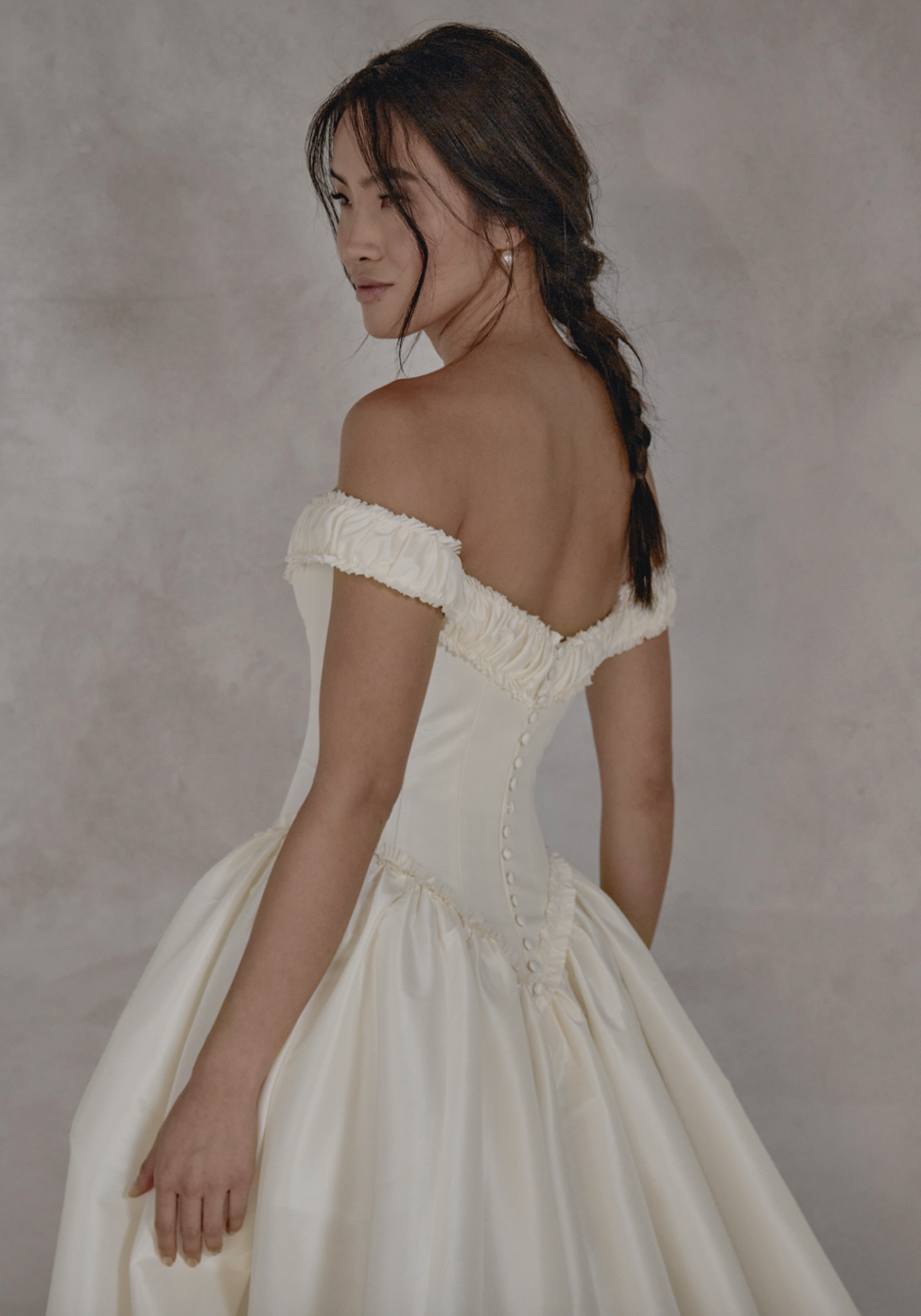 Woman wearing an off-the-shoulder white dress with a fitted bodice and full skirt, posing with her back turned slightly, looking over her shoulder, with brown hair styled in a loose braid against a neutral background.
