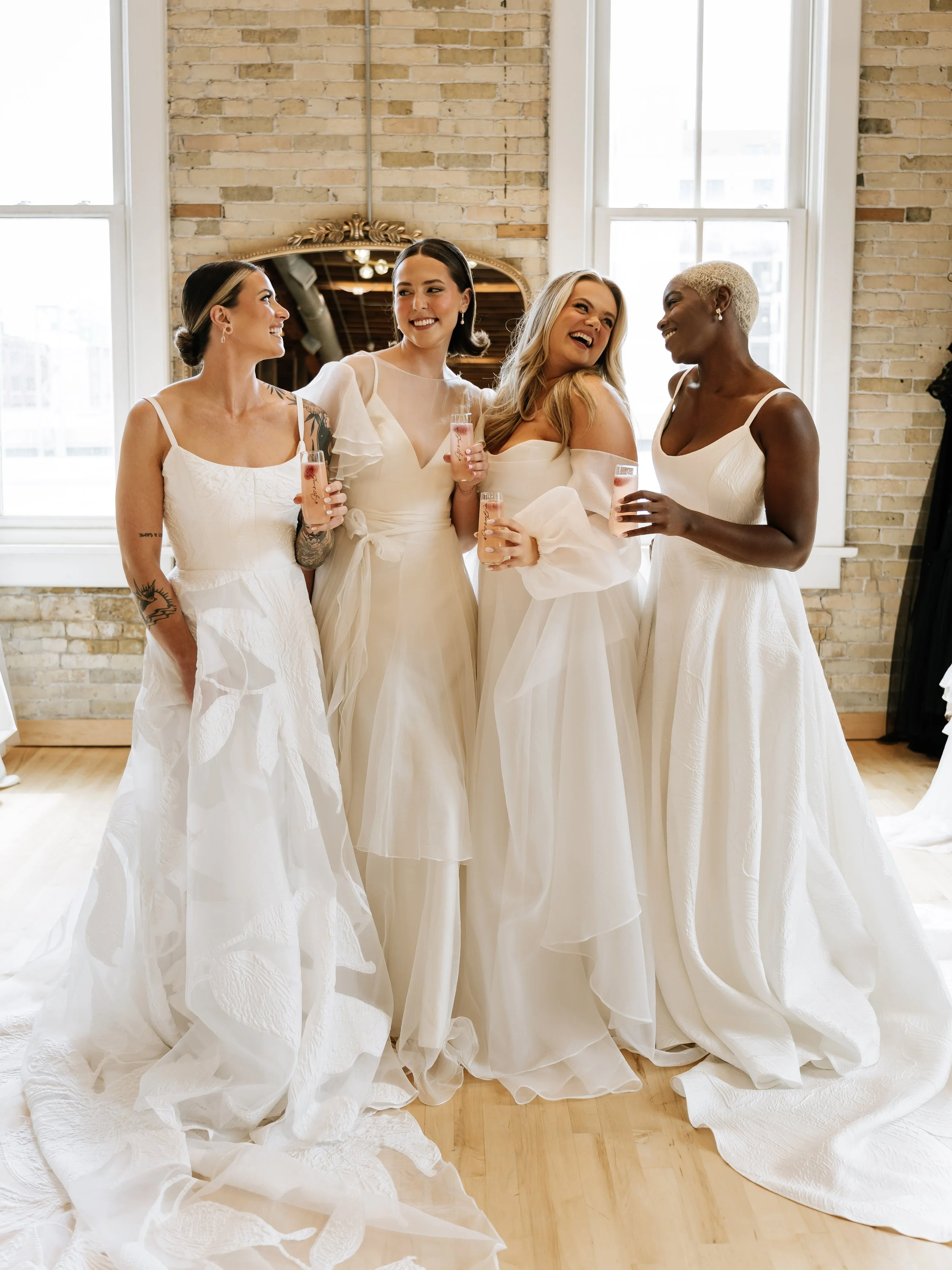 Four women in wedding dresses holding champagne glasses, standing in front of a brick wall with large windows, smiling and chatting.