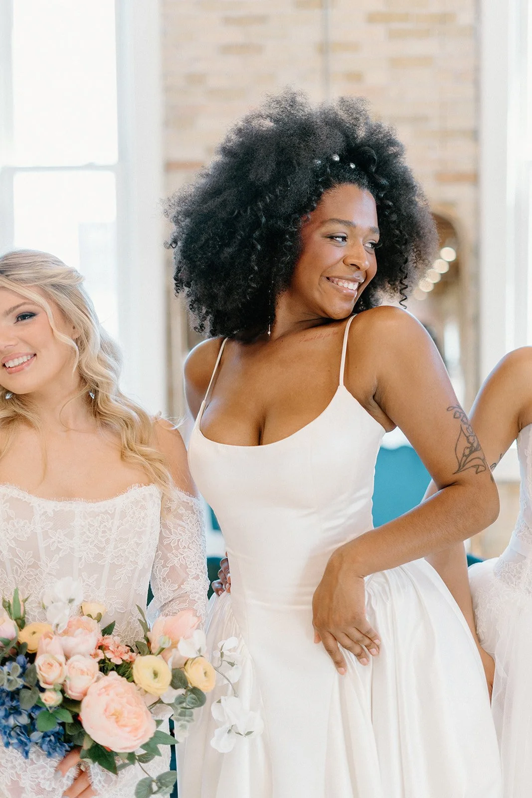 A woman in a white dress with spaghetti straps smiling and posing with her hand on her hip, surrounded by other women in white dresses, one holding a bouquet of flowers, in a bright indoor setting.