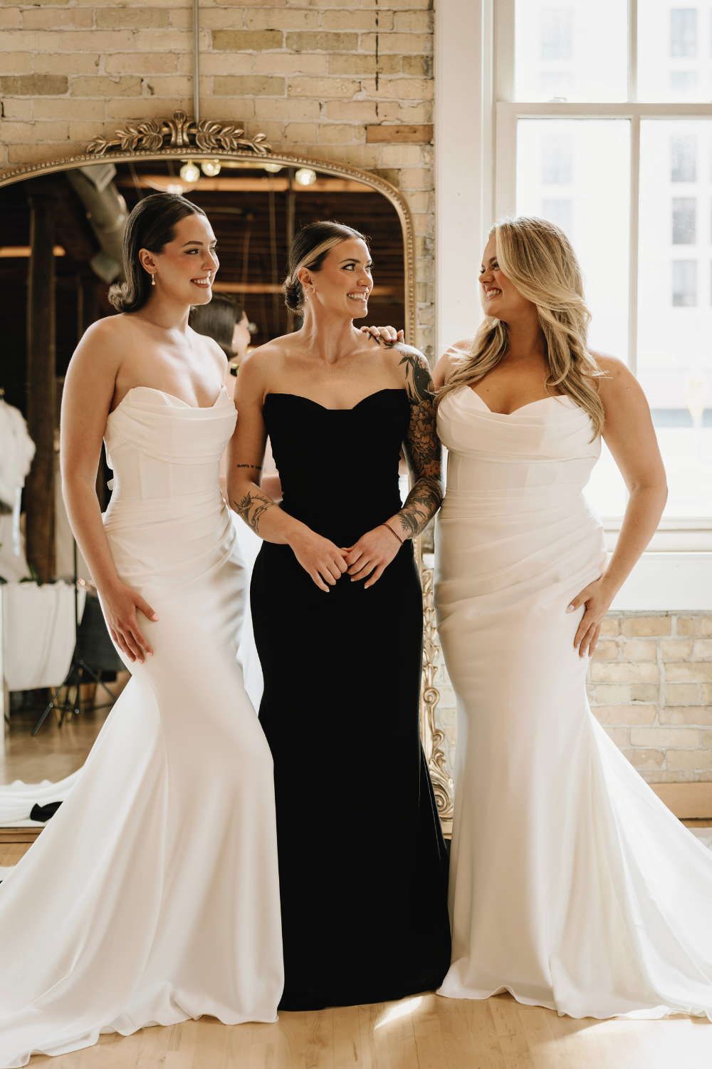 Three women in wedding dresses smiling and looking at each other in a bright, industrial-style room with exposed brick wall and large window.