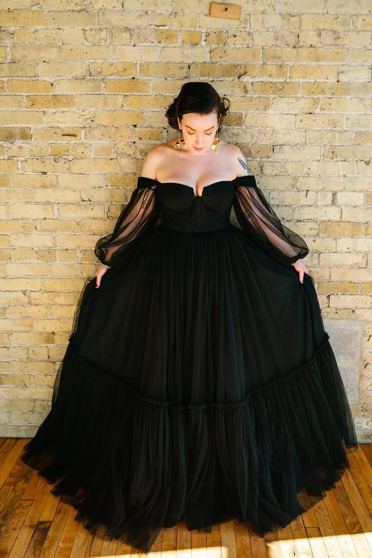 Woman in a black off-shoulder gown with sheer long sleeves, standing against a yellow brick wall.
