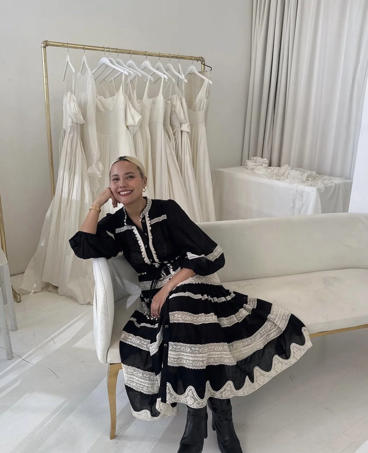A woman smiling and sitting on a white chair in front of a rack of white dresses. The dresses are hanging on hangers in a room with white walls and curtains.