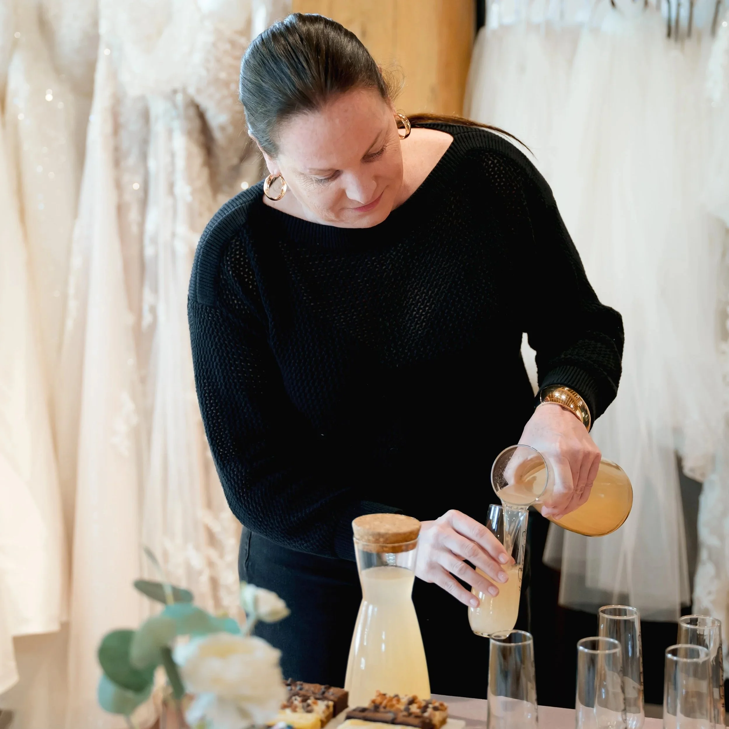 A woman with dark hair in a bun, wearing hoop earrings and a black knit sweater, pouring a drink into a glass at a table with desserts and empty glasses, with a background of lace and fabric decor.