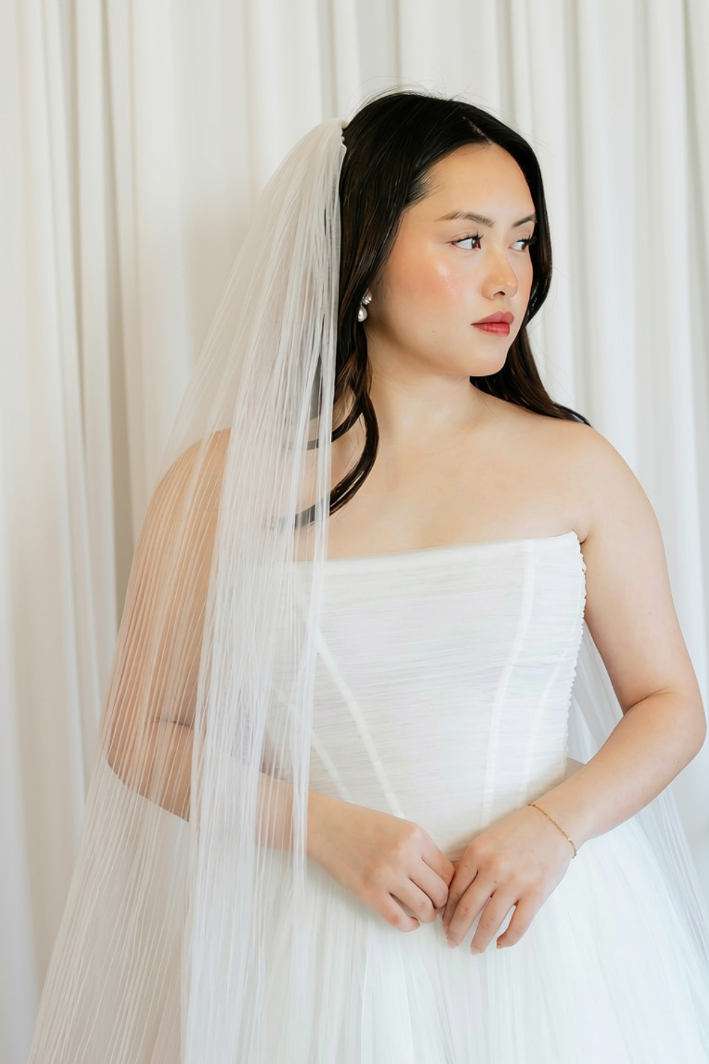 A bride with long dark hair wearing a white strapless wedding dress, pearl earrings, and a veil, standing in front of white curtains.
