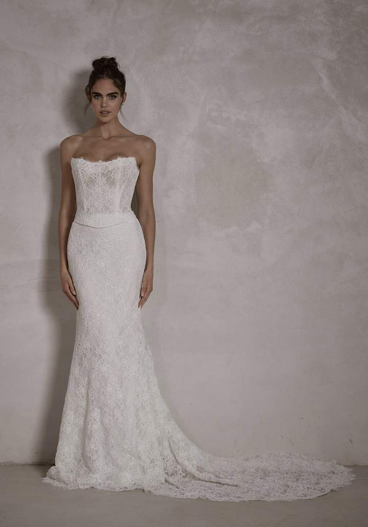 A woman wearing an elegant, strapless white lace wedding dress with a long train, standing against a plain, textured wall.