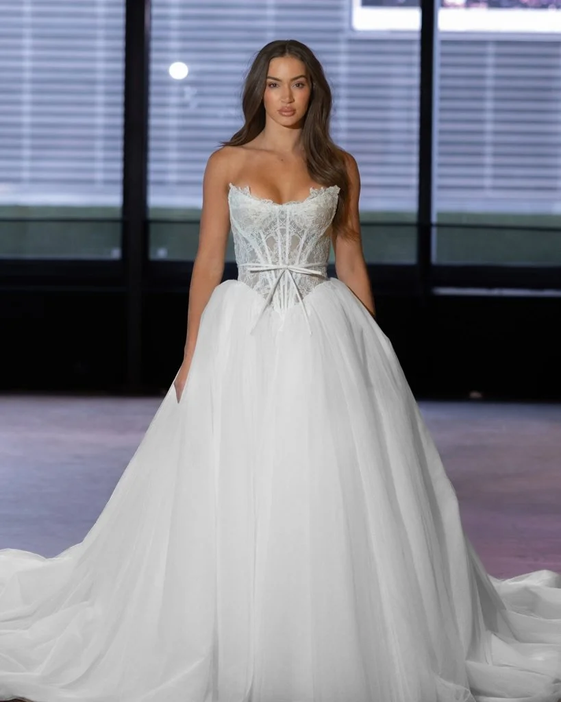 Woman wearing a white wedding gown with lace details on the bodice, standing indoors with large windows and blinds in the background.
