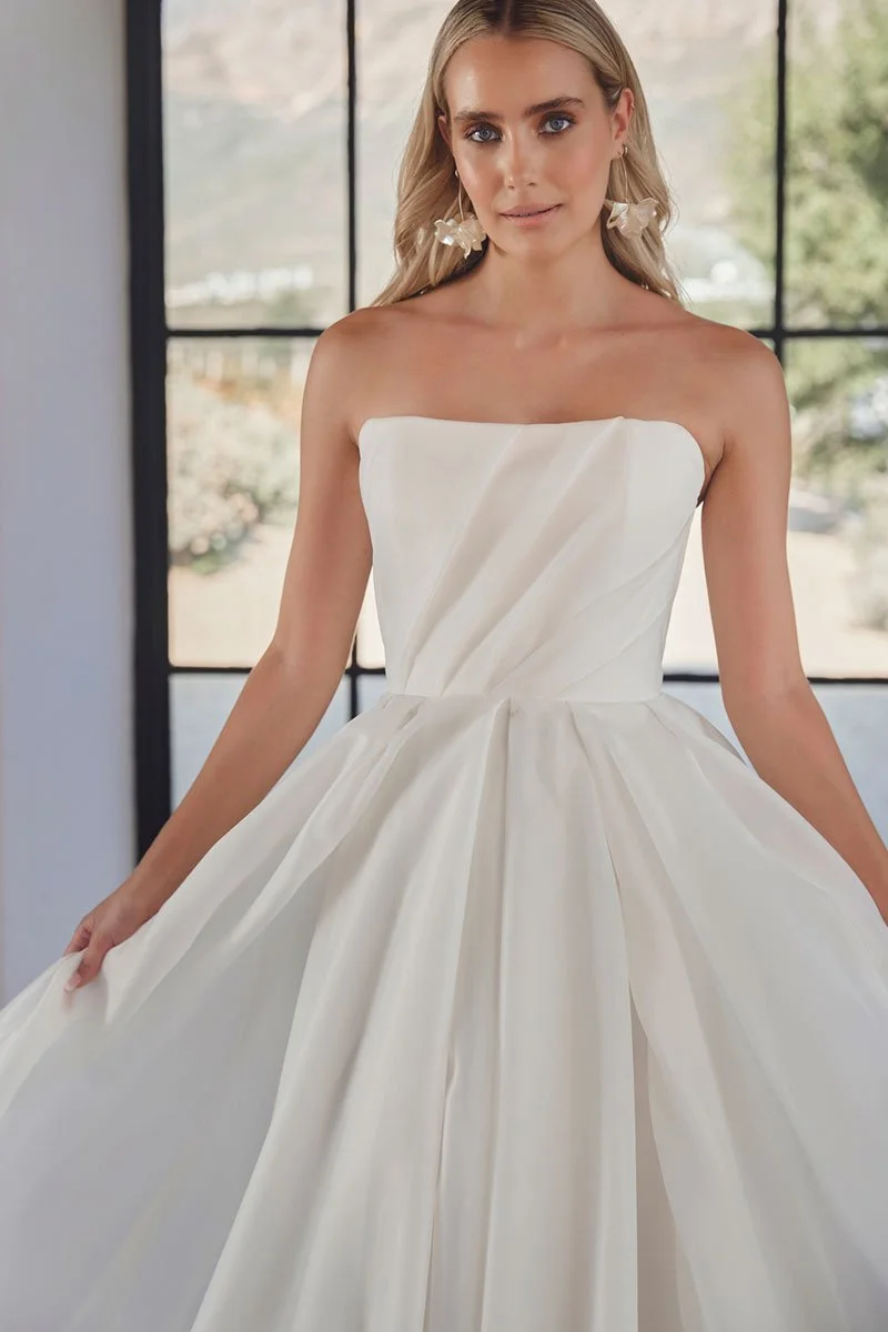 A woman in a strapless white wedding gown with a full skirt standing indoors with large windows in the background.