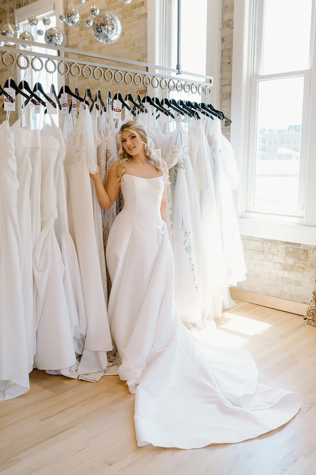 A woman in a wedding dress trying on a gown in a bridal shop with white dresses hanging on a rack, large windows, and a brick wall.