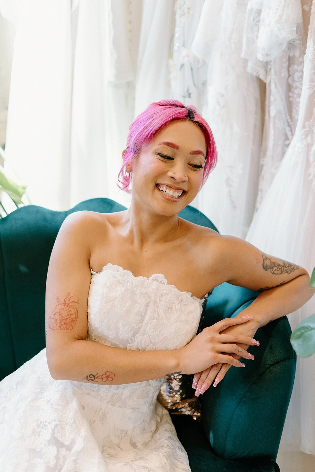 A woman with pink hair, wearing a white lace strapless dress, smiling and sitting on a green velvet chair in front of white lace curtains and wedding dresses.