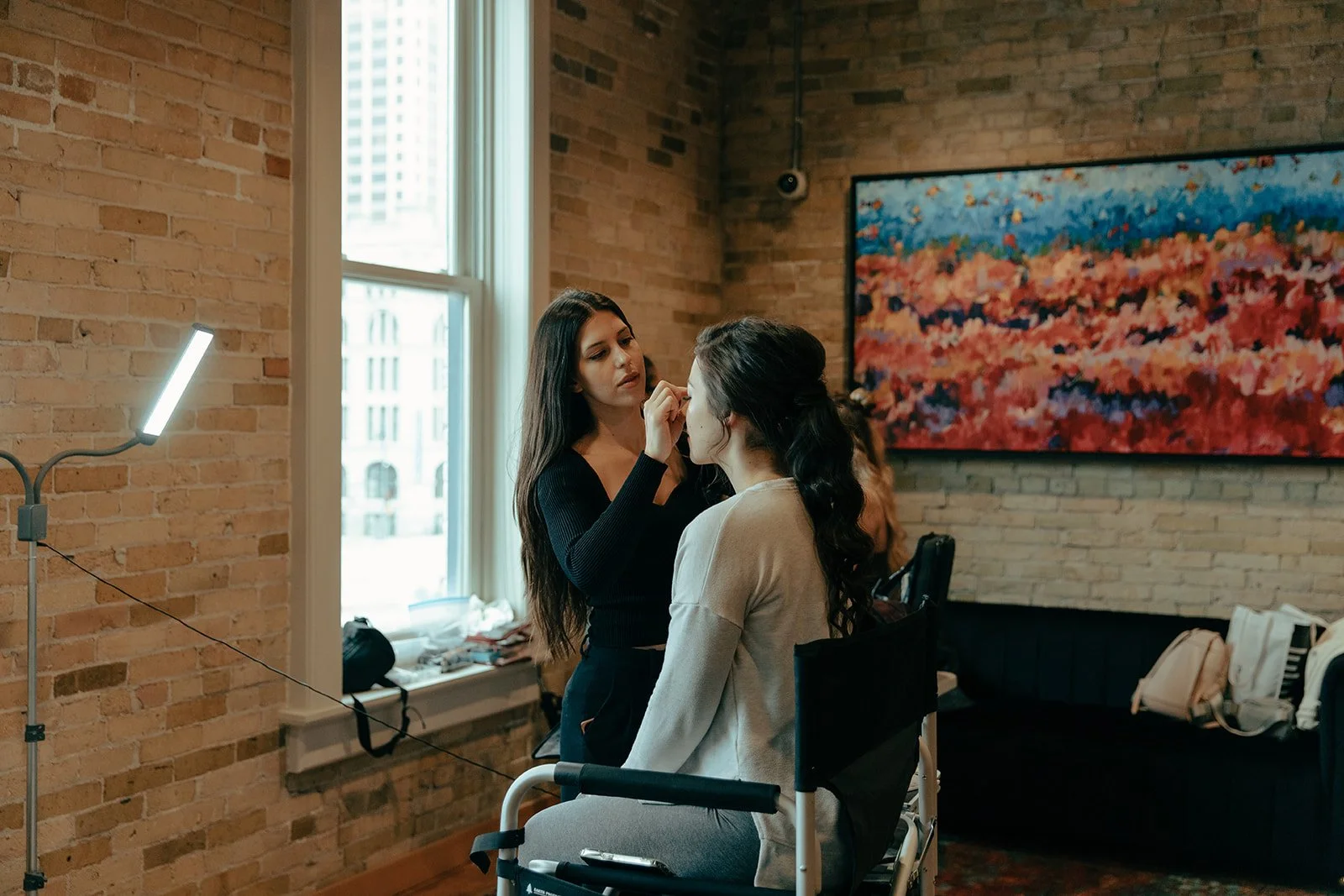 A makeup artist applying makeup on a woman sitting in a chair in a room with exposed brick walls and a large colorful painting. The woman in the chair has long dark hair and is wearing a light-colored sweater. There is a window on the left letting in natural light, and a table with makeup supplies in front of the window.
