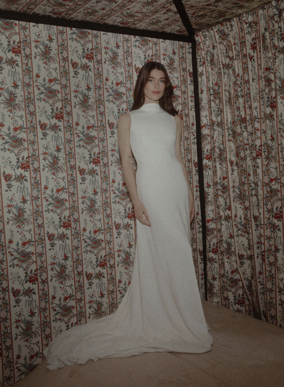A woman in a white, sleeveless, high-neck gown with a long train, standing in a corner with floral-patterned wallpaper.