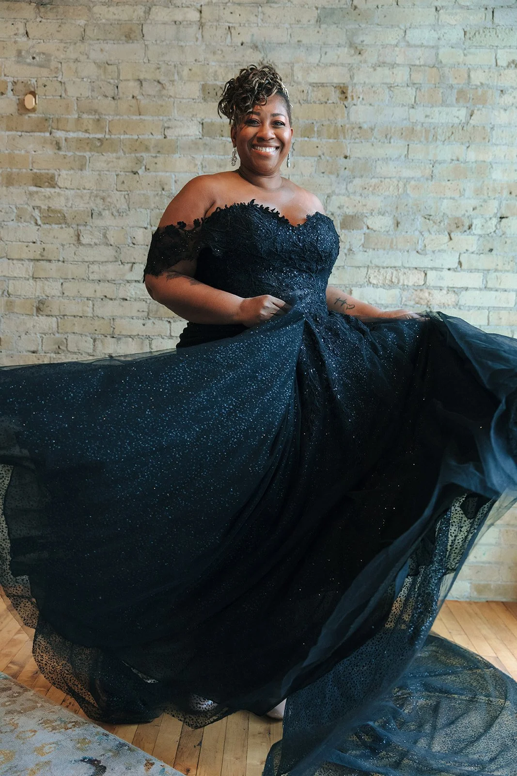A woman wearing a black evening gown with off-the-shoulder sleeves, lace, and glitter, is smiling and holding the skirt of her dress. She has short curly hair and is standing against a brick wall, indoors.