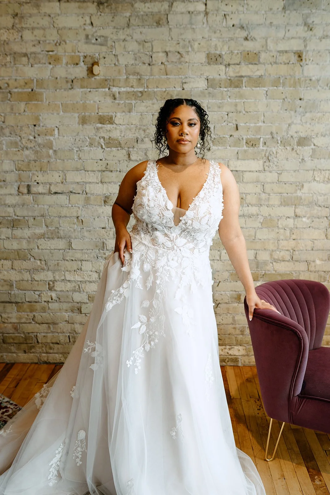 A woman dressed in a white wedding gown with lace and floral details, posing in front of a beige brick wall, with one hand on a burgundy velvet armchair.