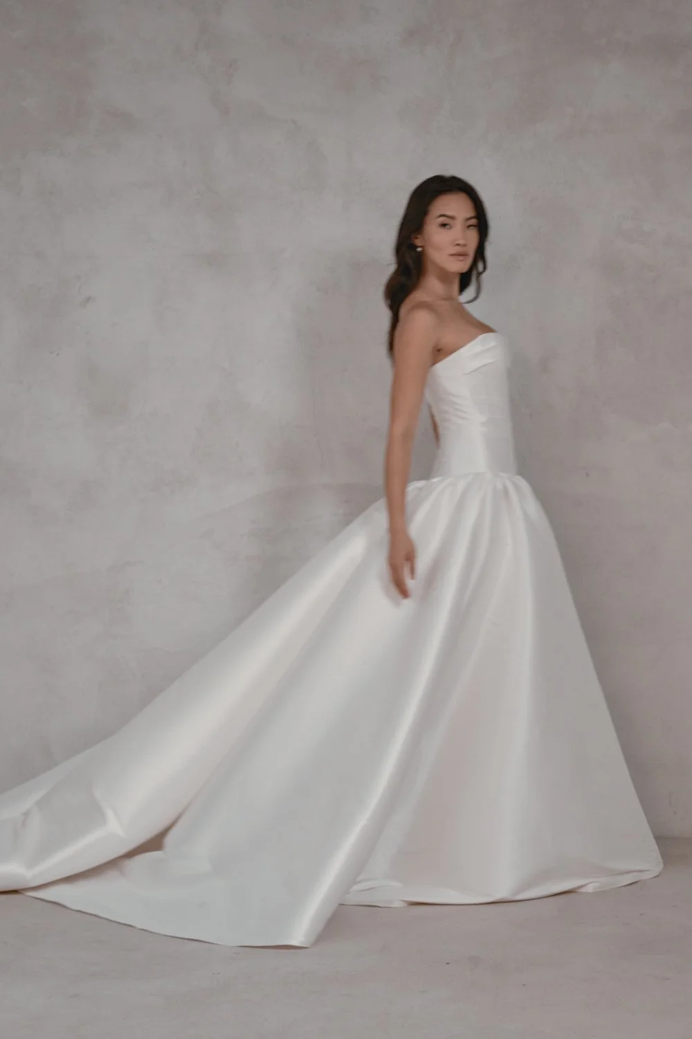 A woman in a strapless white wedding gown with a full skirt and train, standing against a textured gray wall.