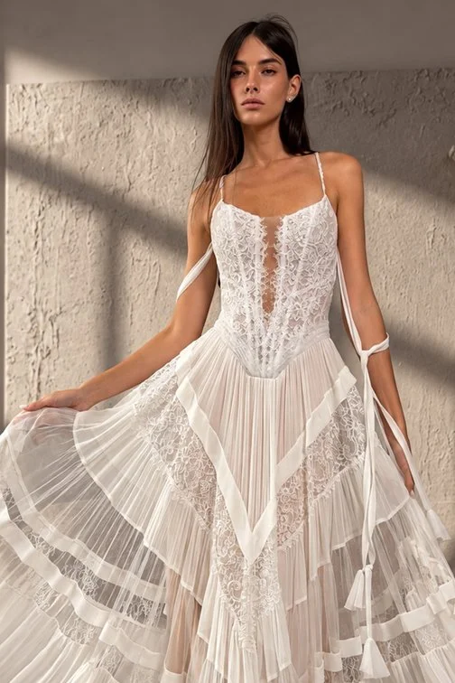 A woman in a white lace and pleated dress with spaghetti straps posing against a textured wall.