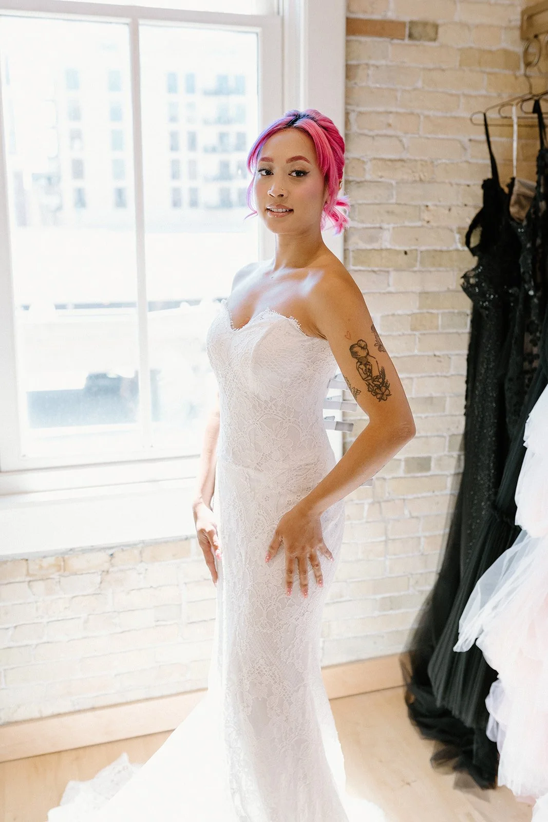 A woman with pink hair wearing a white lace wedding dress standing in a bright room with a brick wall and a large window.