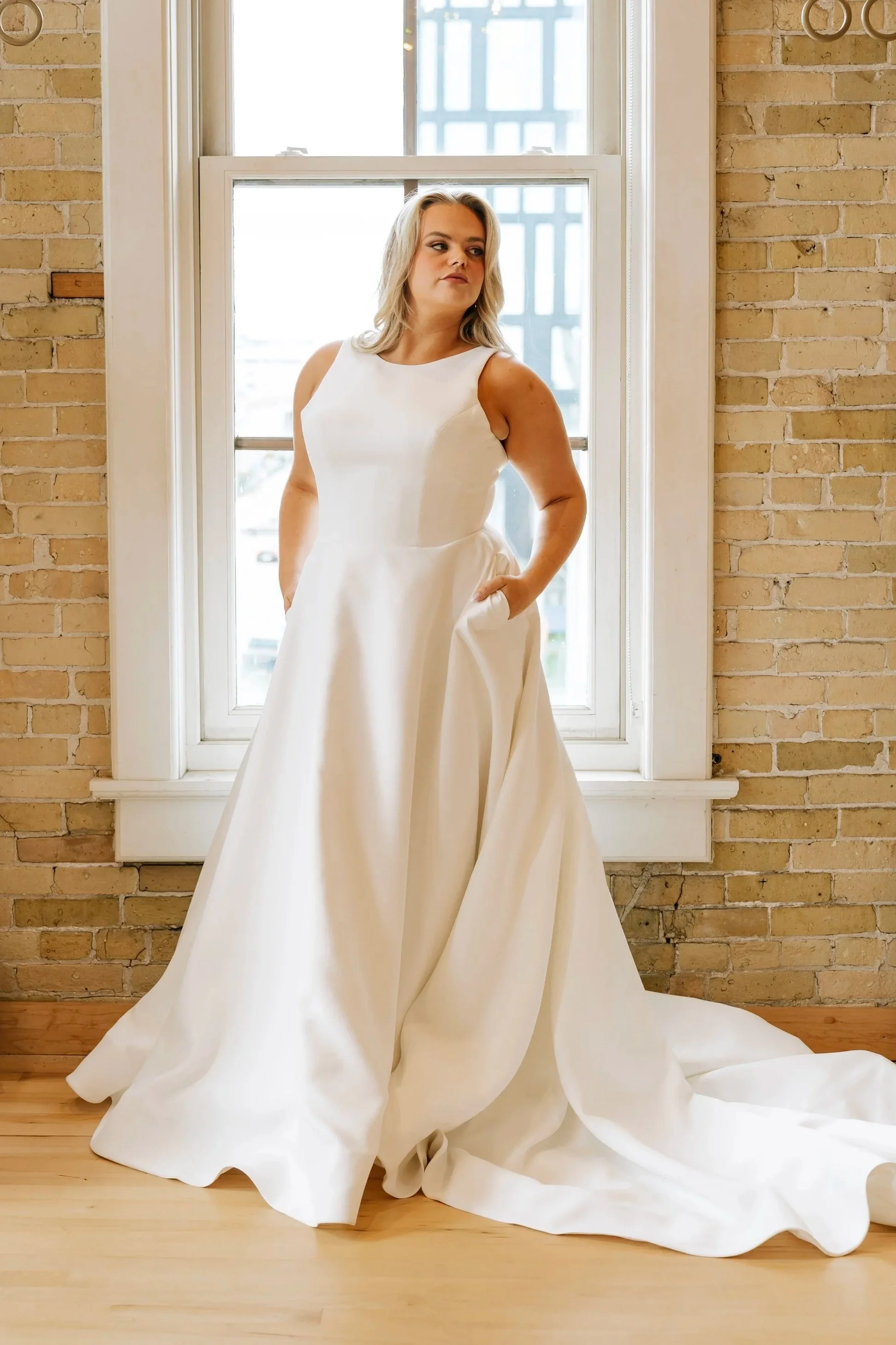 A woman in a white wedding dress stands in front of a window with brick walls on either side. The dress has a sleeveless design and a full skirt that pools on the floor.