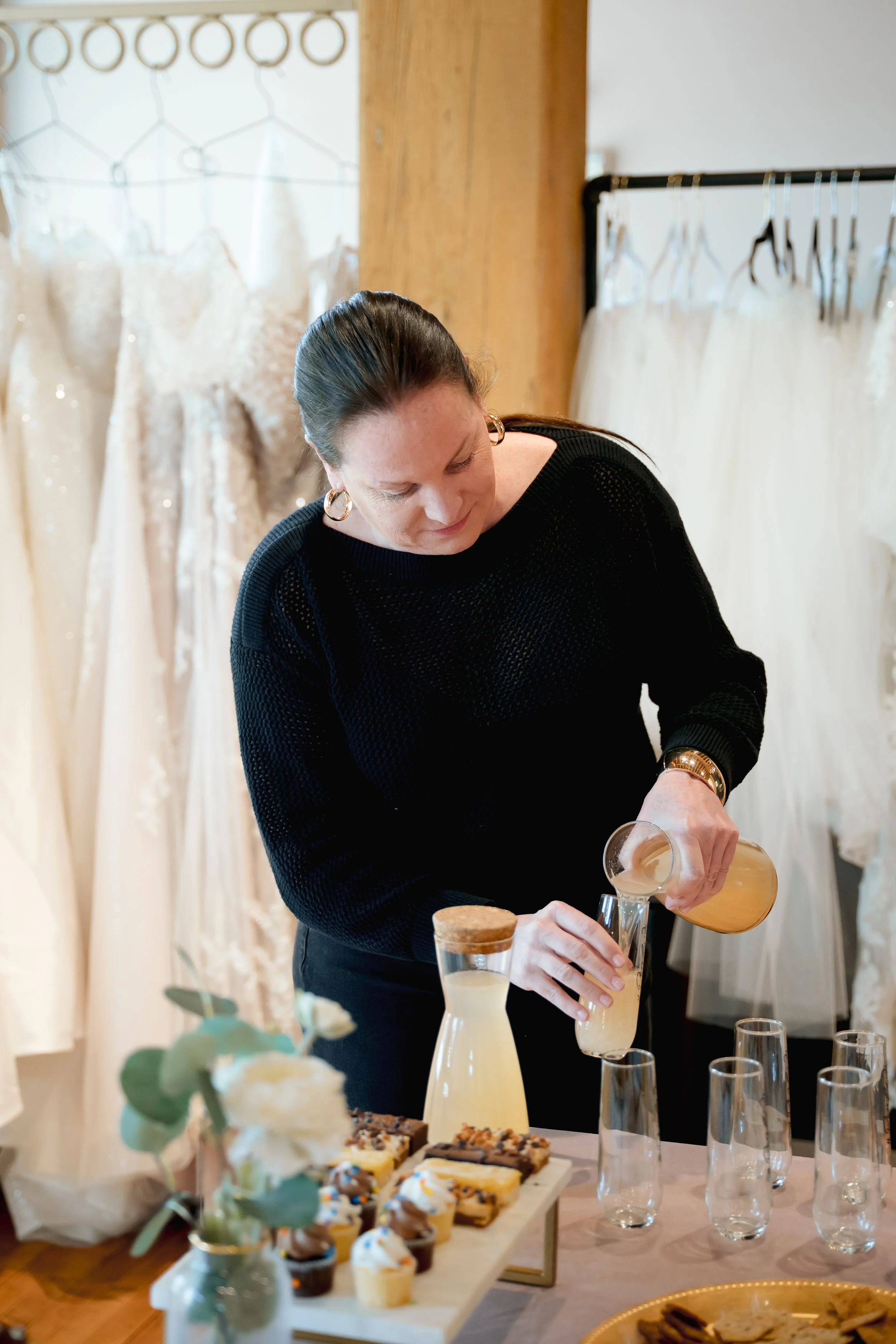 A woman in a black sweater serving drinks at a gathering with desserts and refreshments on a table, with dresses hanging on racks in the background.