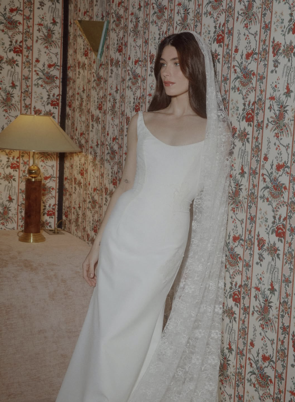A woman in a white wedding dress with lace veil standing indoors against a floral patterned wallpaper background, near a table with a lamp.