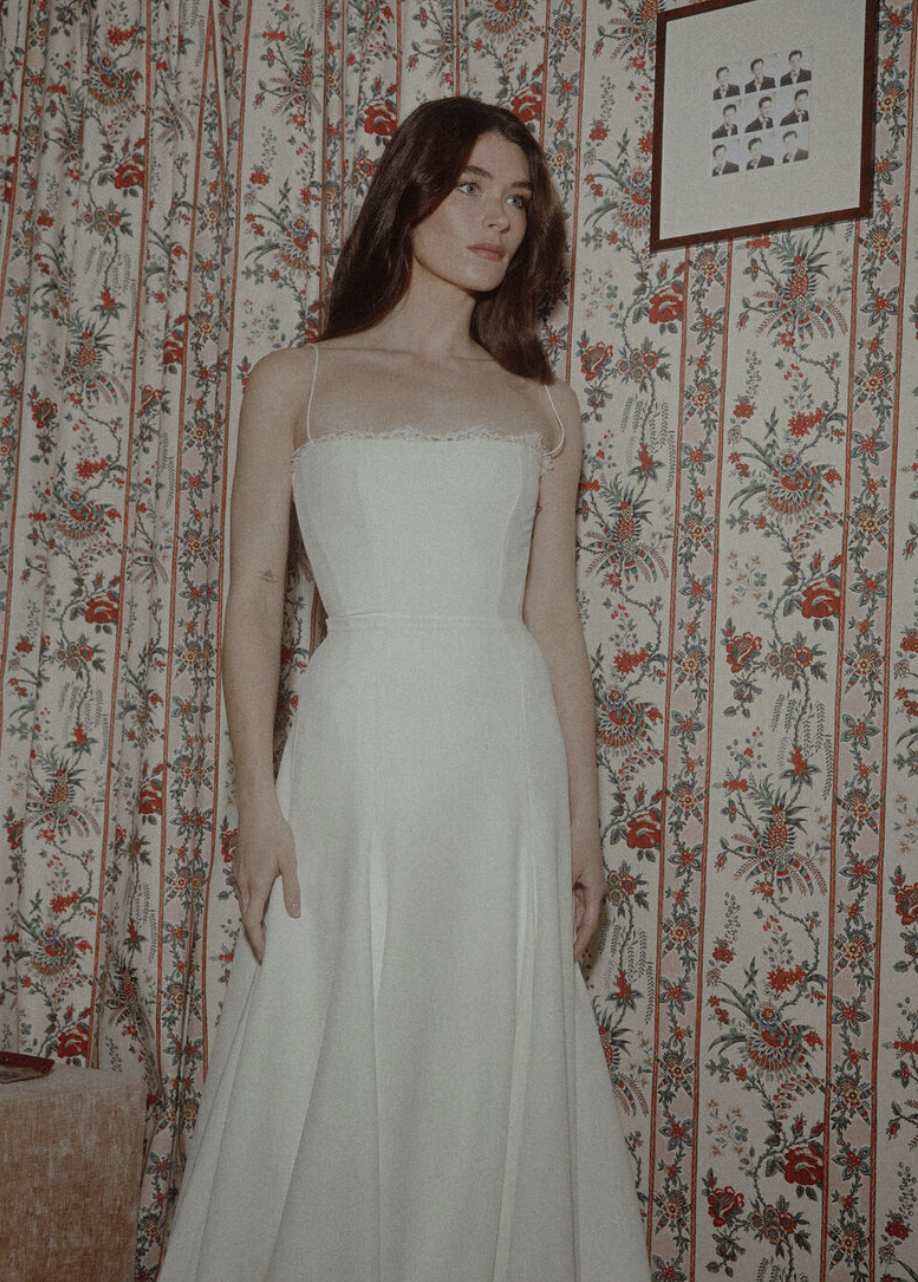 A woman with long brown hair wearing a white dress standing in front of a floral patterned wallpaper. There is a framed photo on the wall behind her.