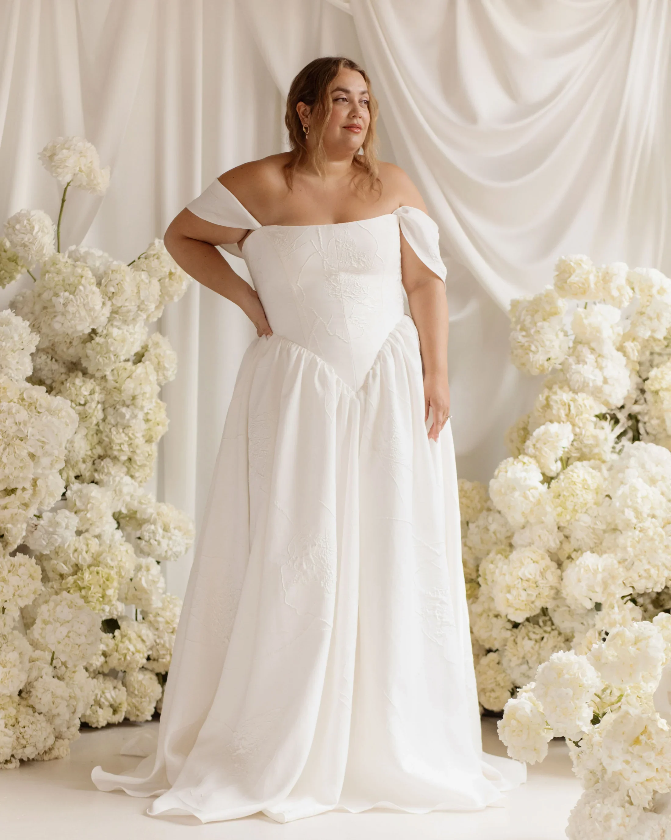 A woman in a white off-shoulder wedding dress standing among white floral arrangements and draped white curtains.