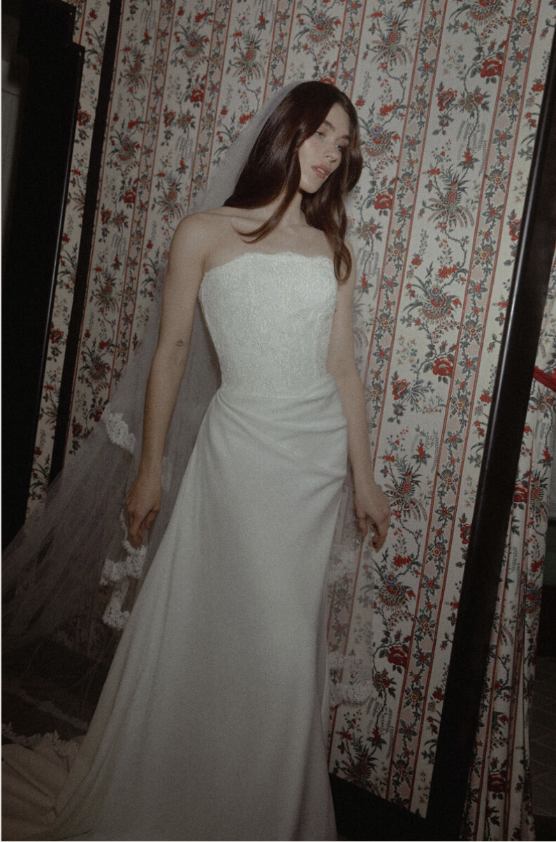 A woman in a white wedding dress with a strapless lace bodice and a veil, standing in front of a patterned wallpaper.