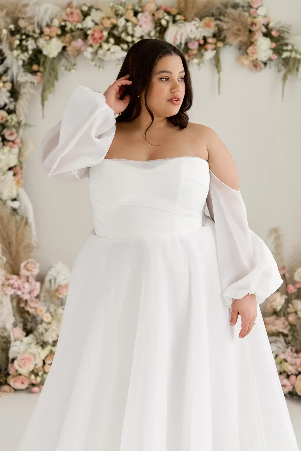 A woman in a white off-shoulder wedding dress with sheer long sleeves, standing in front of a floral arch decor with pink and white flowers.
