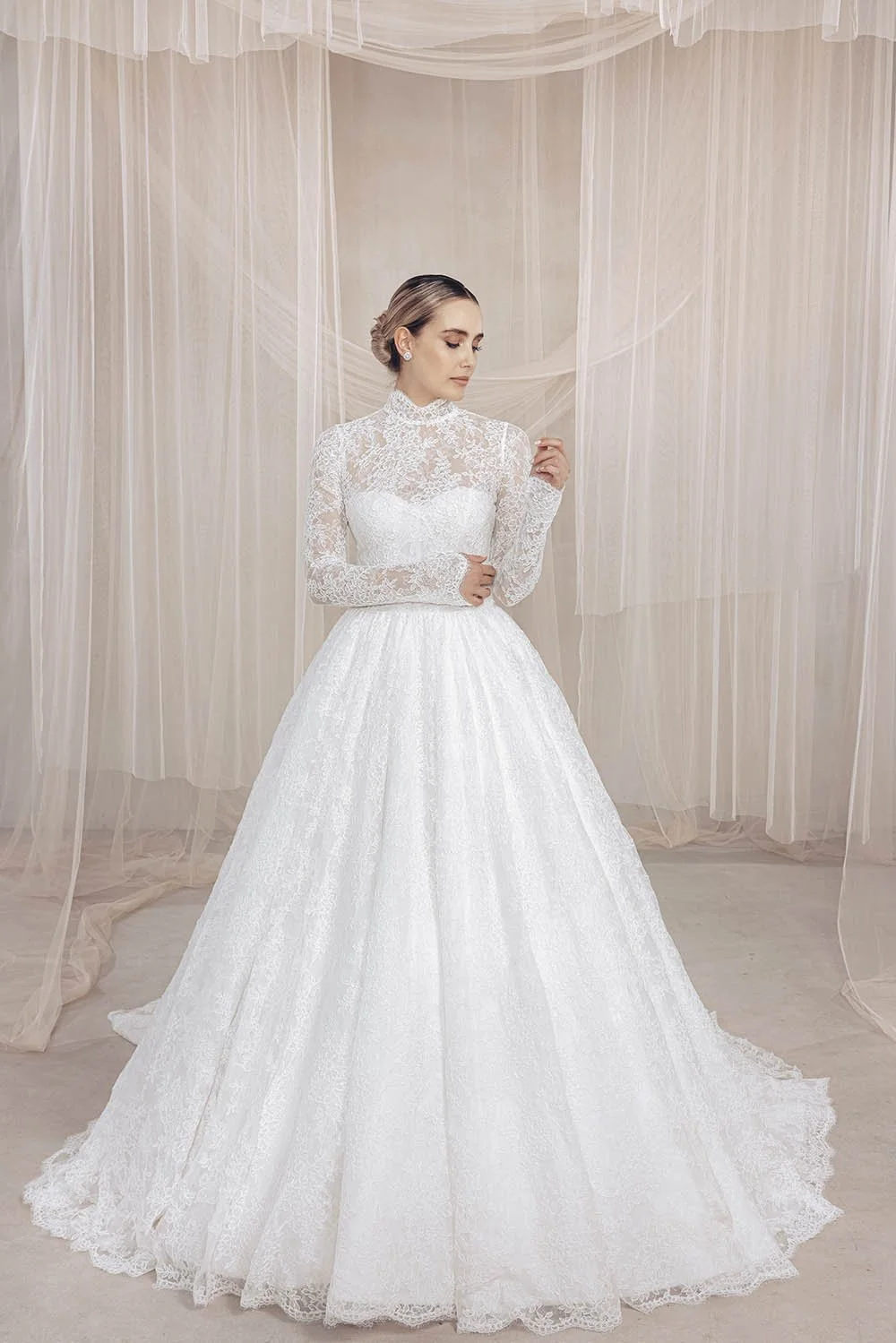A woman in a wedding dress with lace details stands in a softly lit room with sheer curtains.