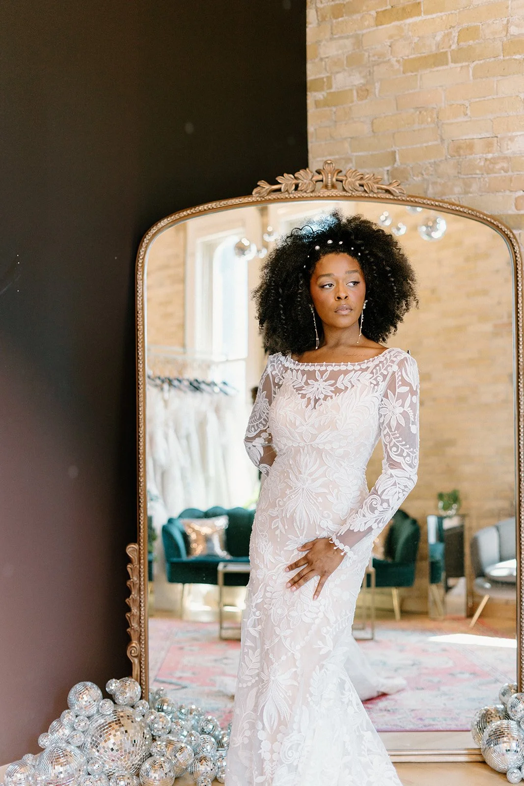 A woman in a white embroidered wedding dress looking into a mirror surrounded by disco balls in a room with exposed brick walls and large windows.